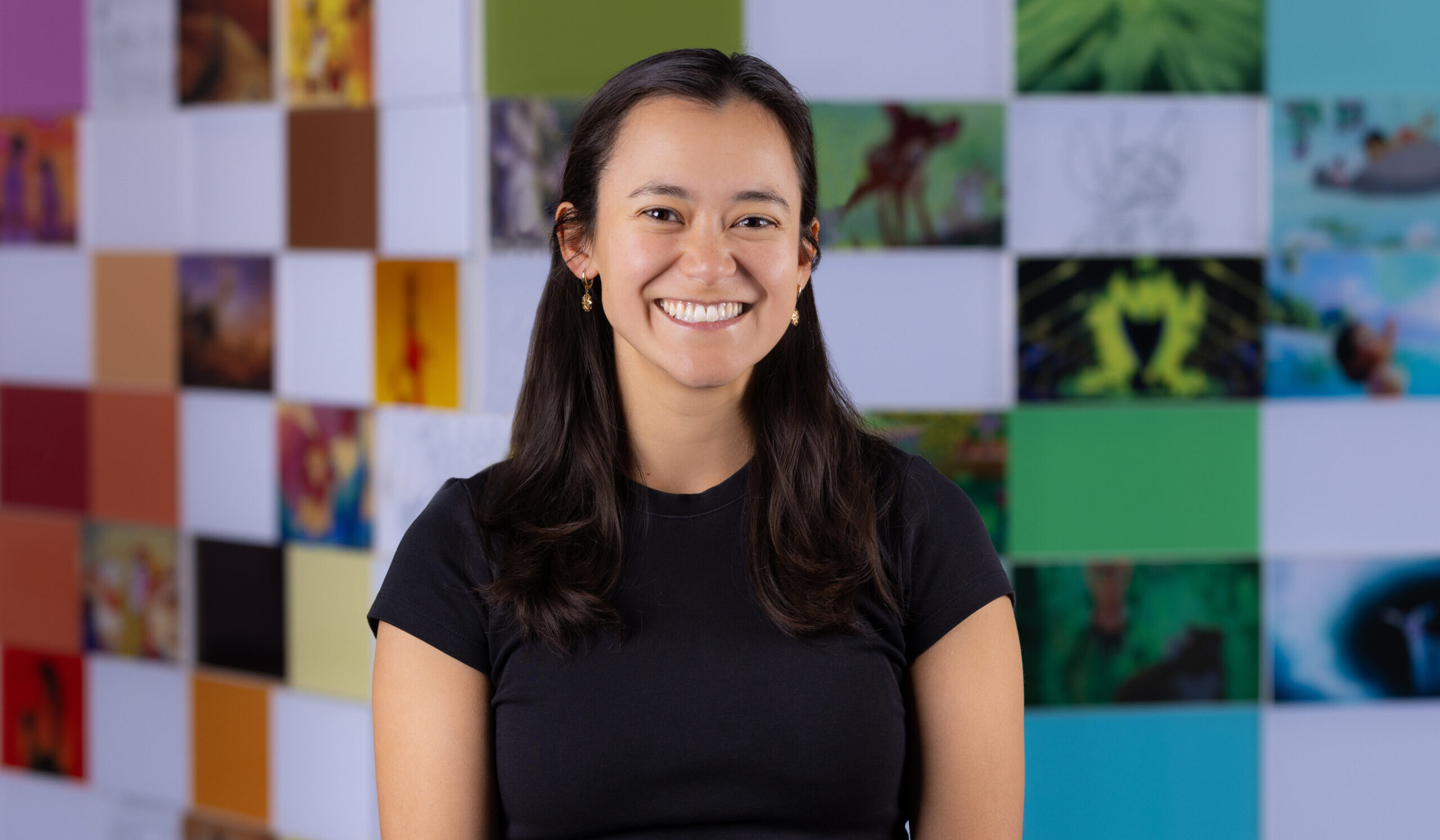 Person in a black shirt smiles in front of a colorful wall covered with small, square images.
