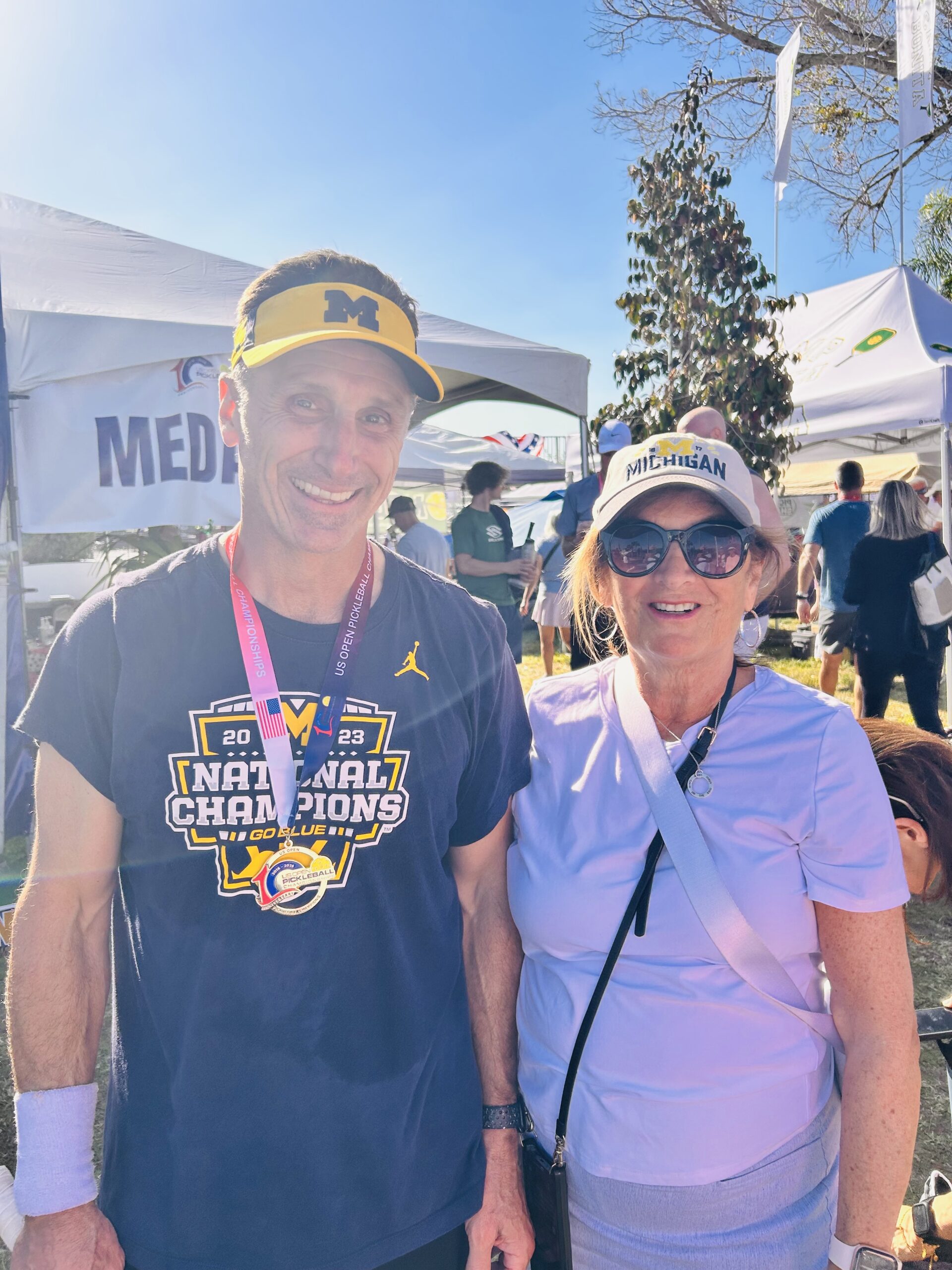 Two people in Michigan apparel stand outdoors at an event, smiling, with tents and other people in the background.