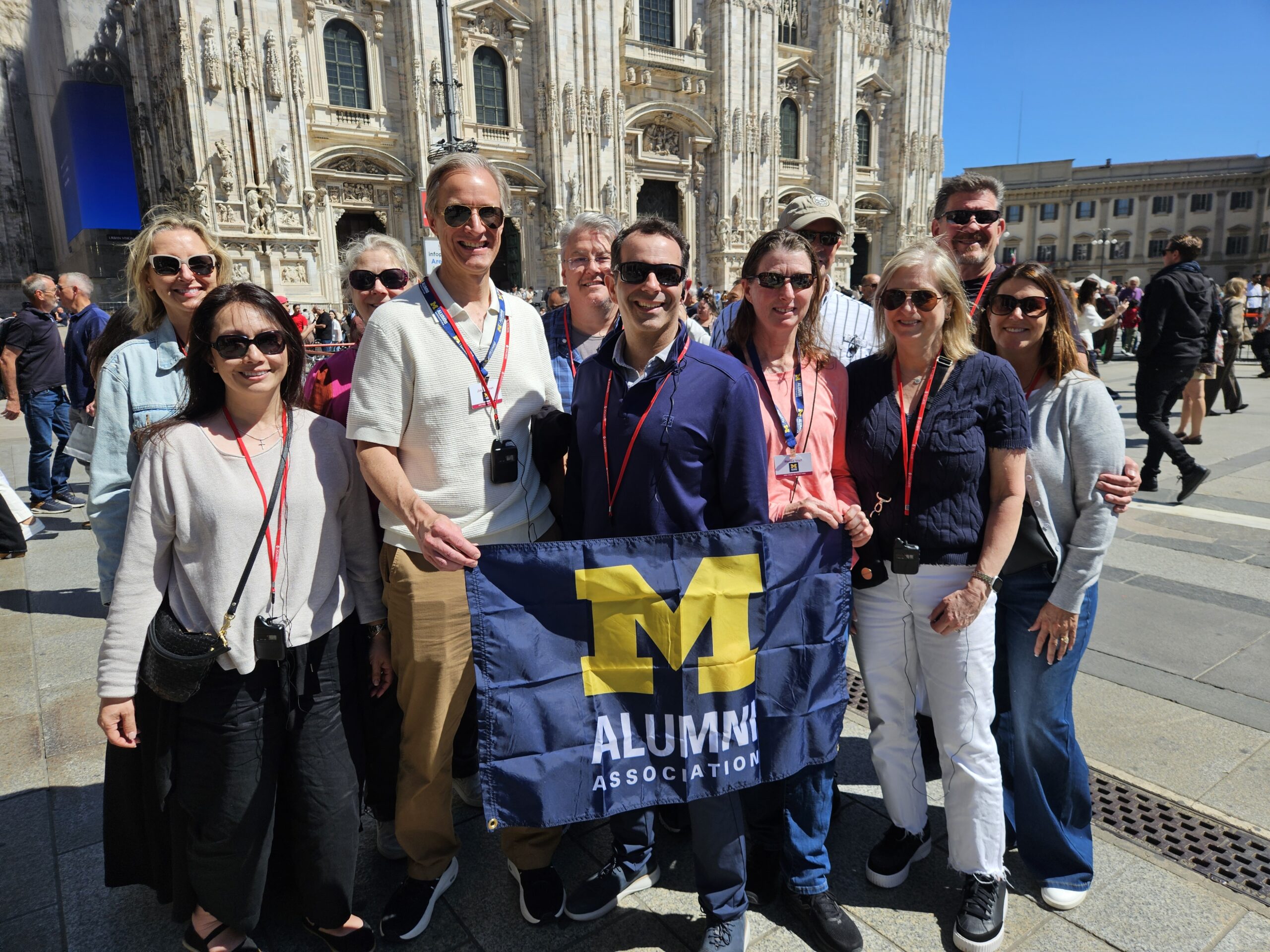 People wearing sunglasses hold a University of Michigan Alumni Association flag in front of a historic building.