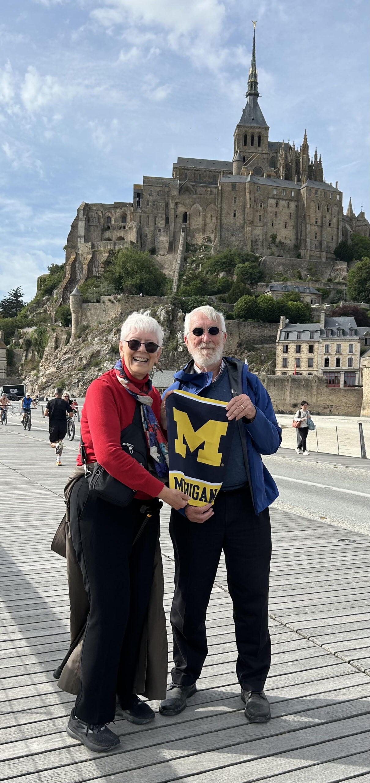 Two people stand outdoors holding a Michigan flag, with a castle-like structure on a hill in the background.