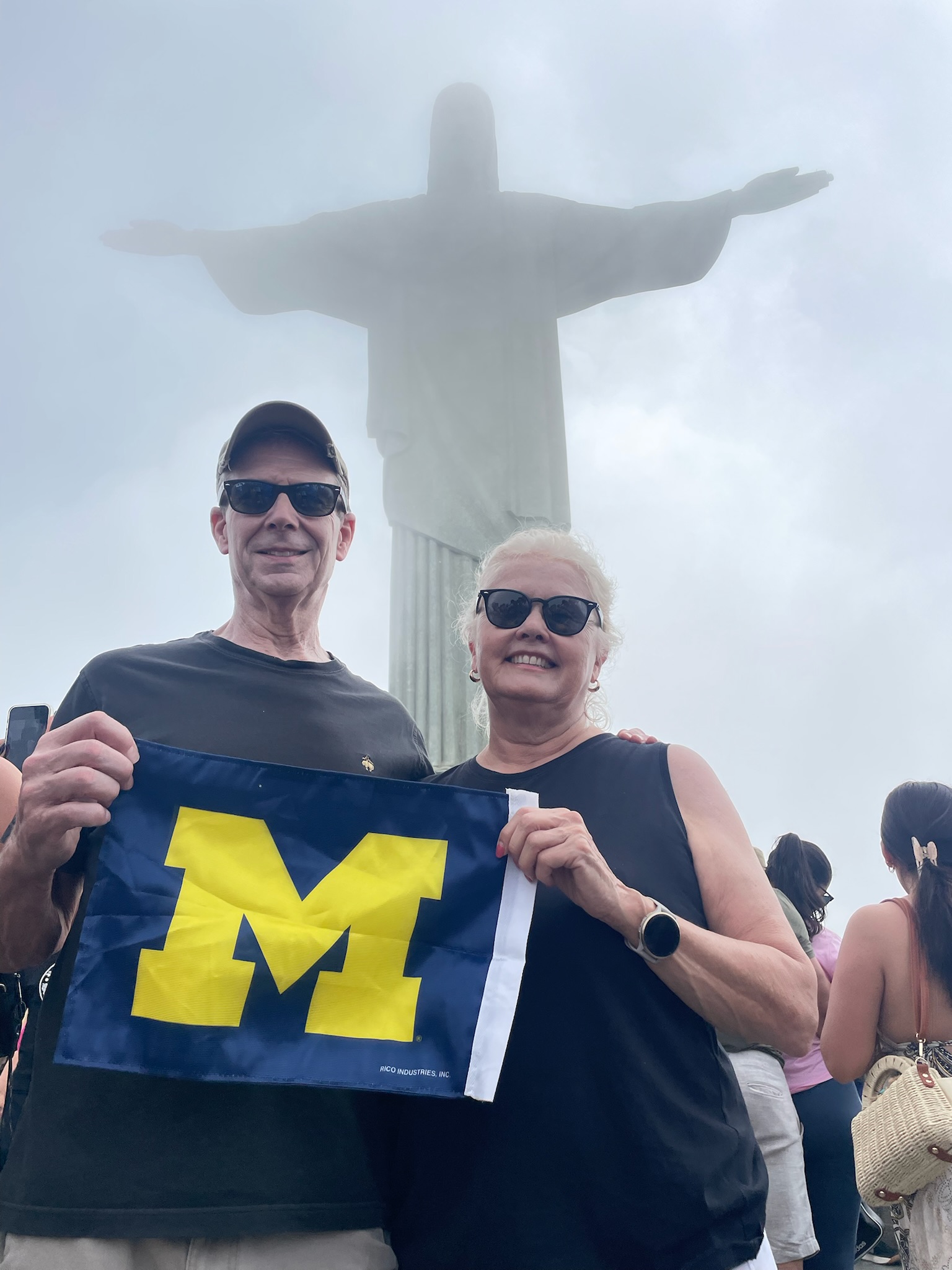 Two people stand in front of the Christ the Redeemer statue on a cloudy day, holding a University of Michigan maize flag.