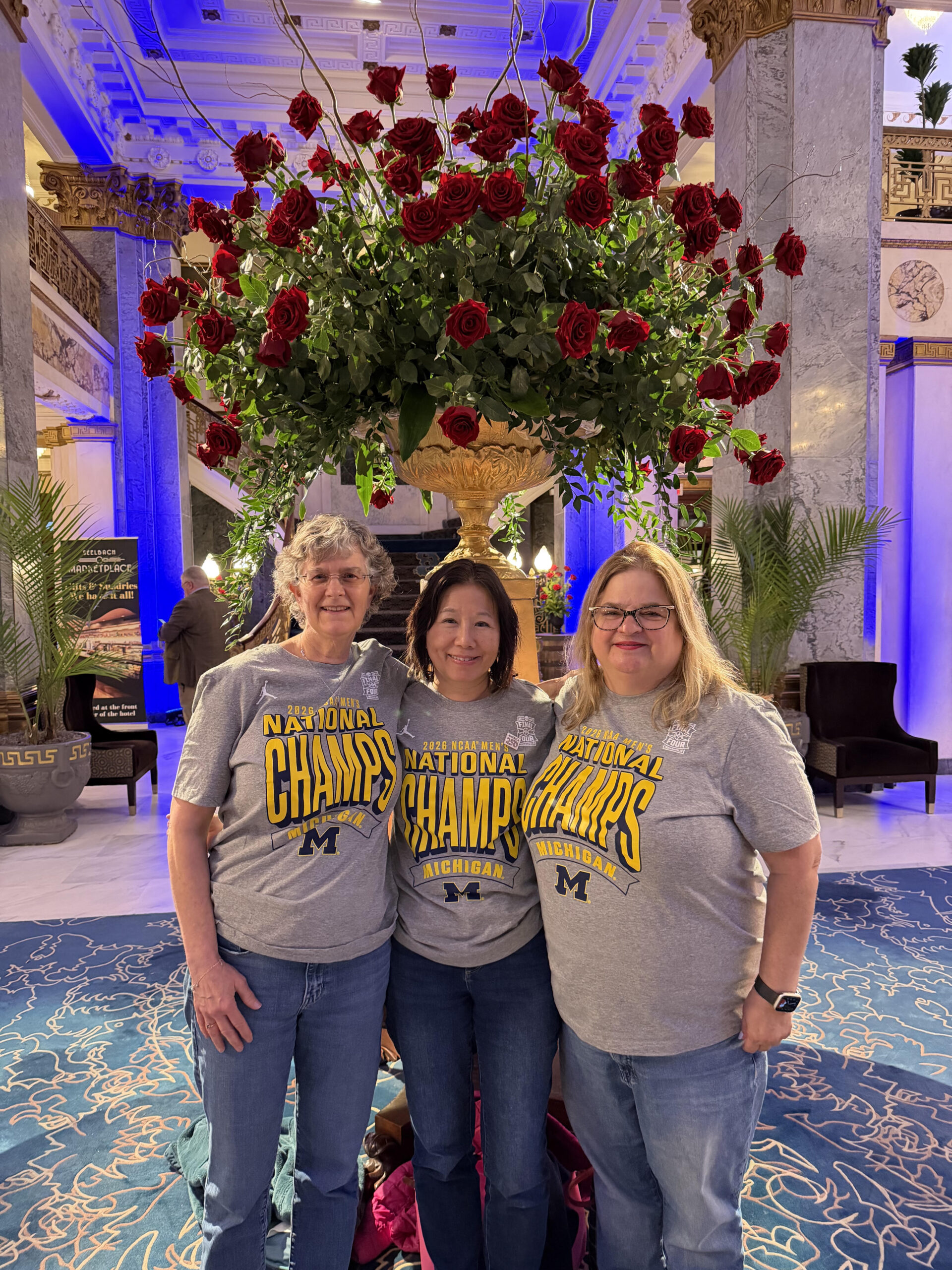Three people in Michigan National Champs shirts stand together in a grand lobby with a large rose arrangement behind them.