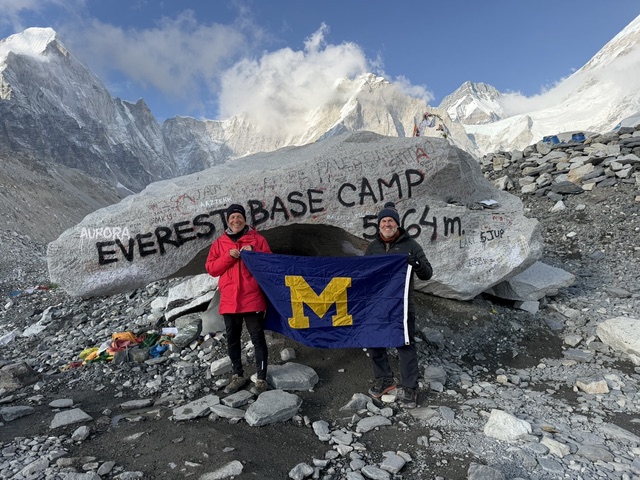 Two people hold a University of Michigan flag in front of the Everest Base Camp 5364m rock sign, with mountains around.