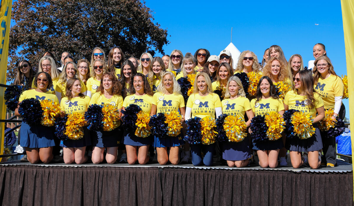 A large group of women pose on a stage. They are all wearing yellow shirts that say Michigan Dance Alumni.