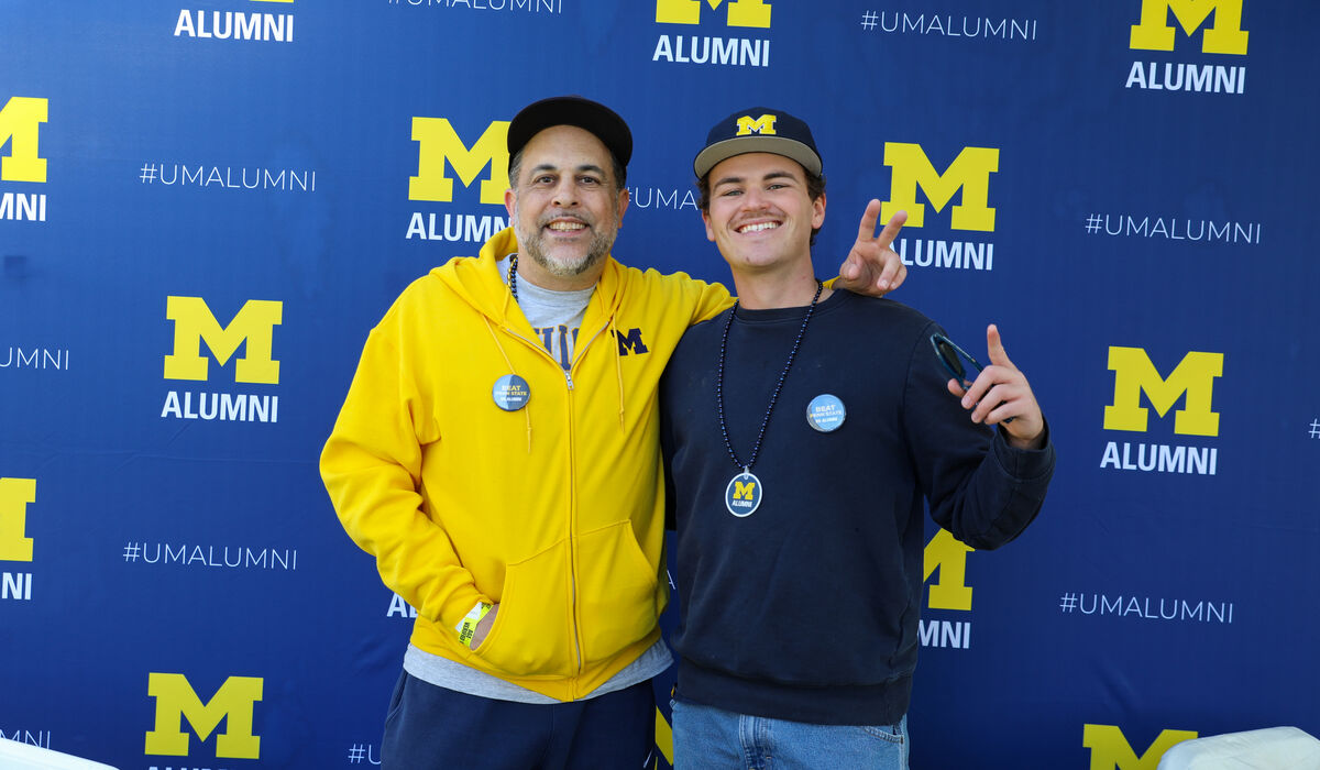 A man in a yellow Michigan shirt smiles with a younger man, who is wearing a navy shirt in front of a backdrop that says M Alumni