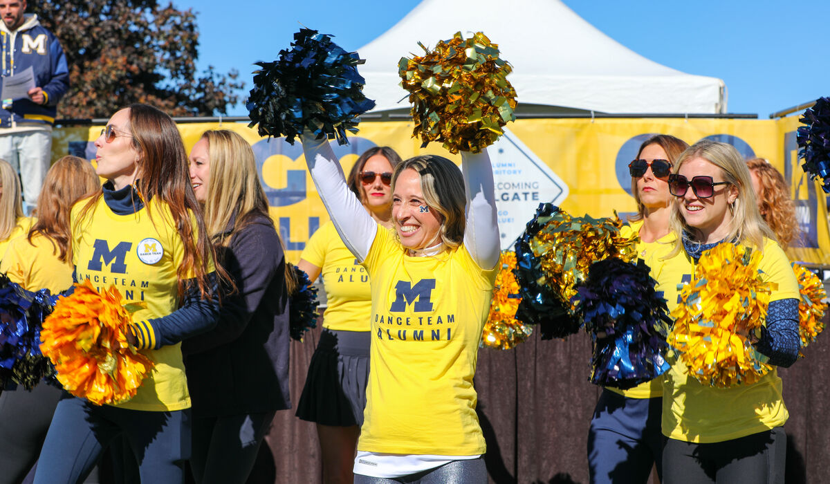A woman wearing a yellow shirt that reads Dance Team Alumni raises her arms