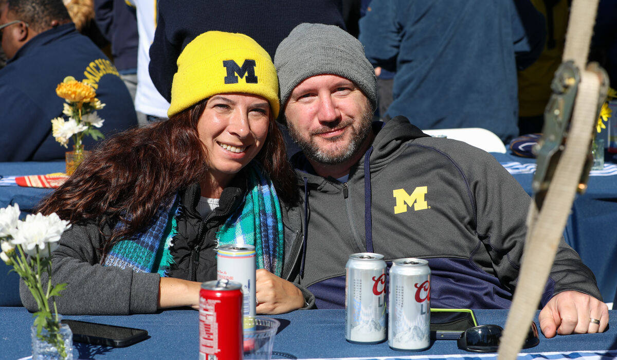 A man and woman sit at a table with drinks on it. They are wearing Michigan clothing.
