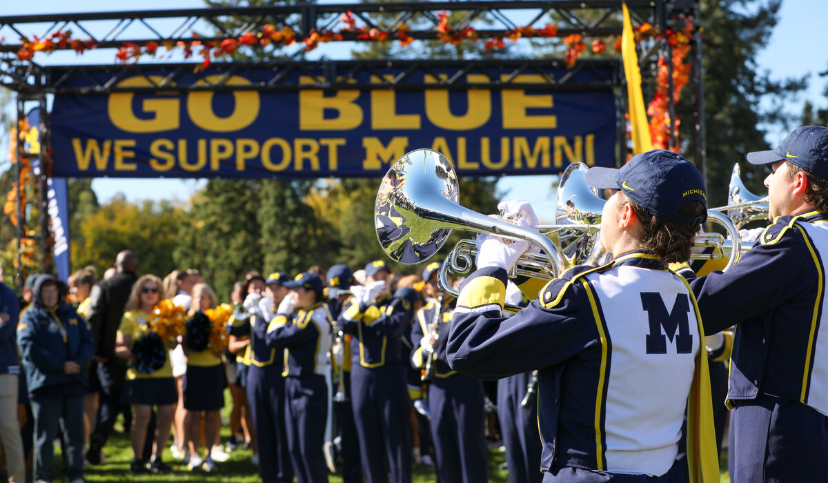 The Michigan marching bad plays in front of a large crowd of people wearing maize and blue in front of a sign that says Go Blue, we support M Alumni