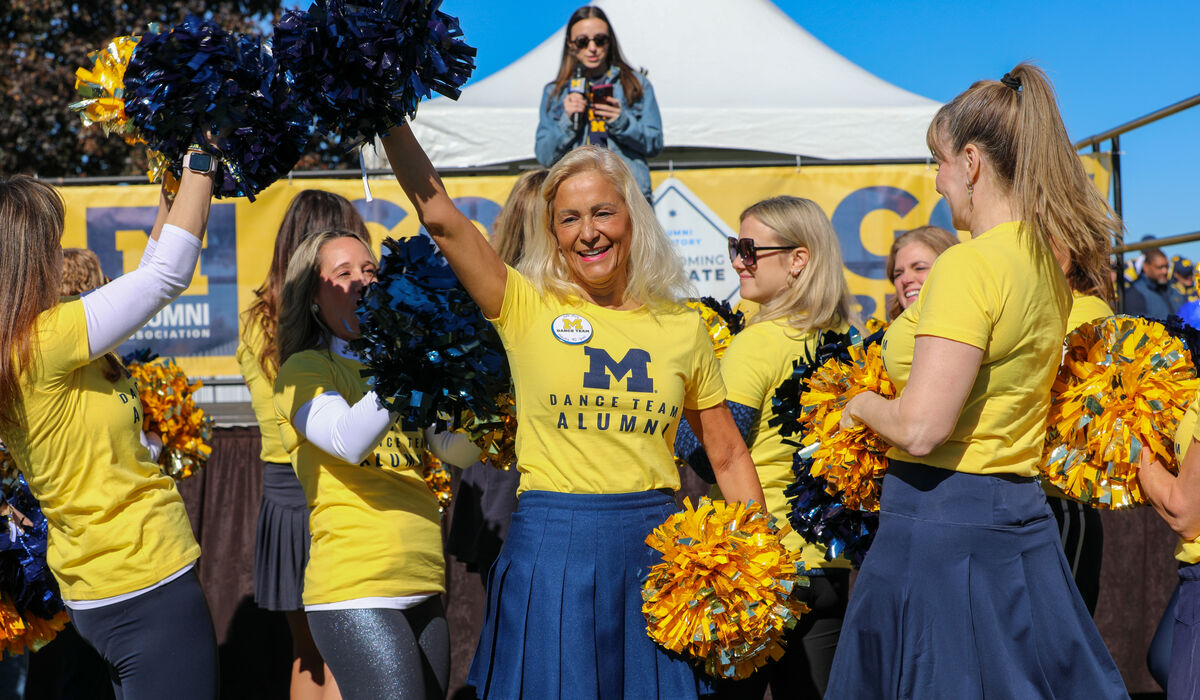 A woman wearing a yellow shirt that says Dance Team Alumni waves to a crowd with a pom pom