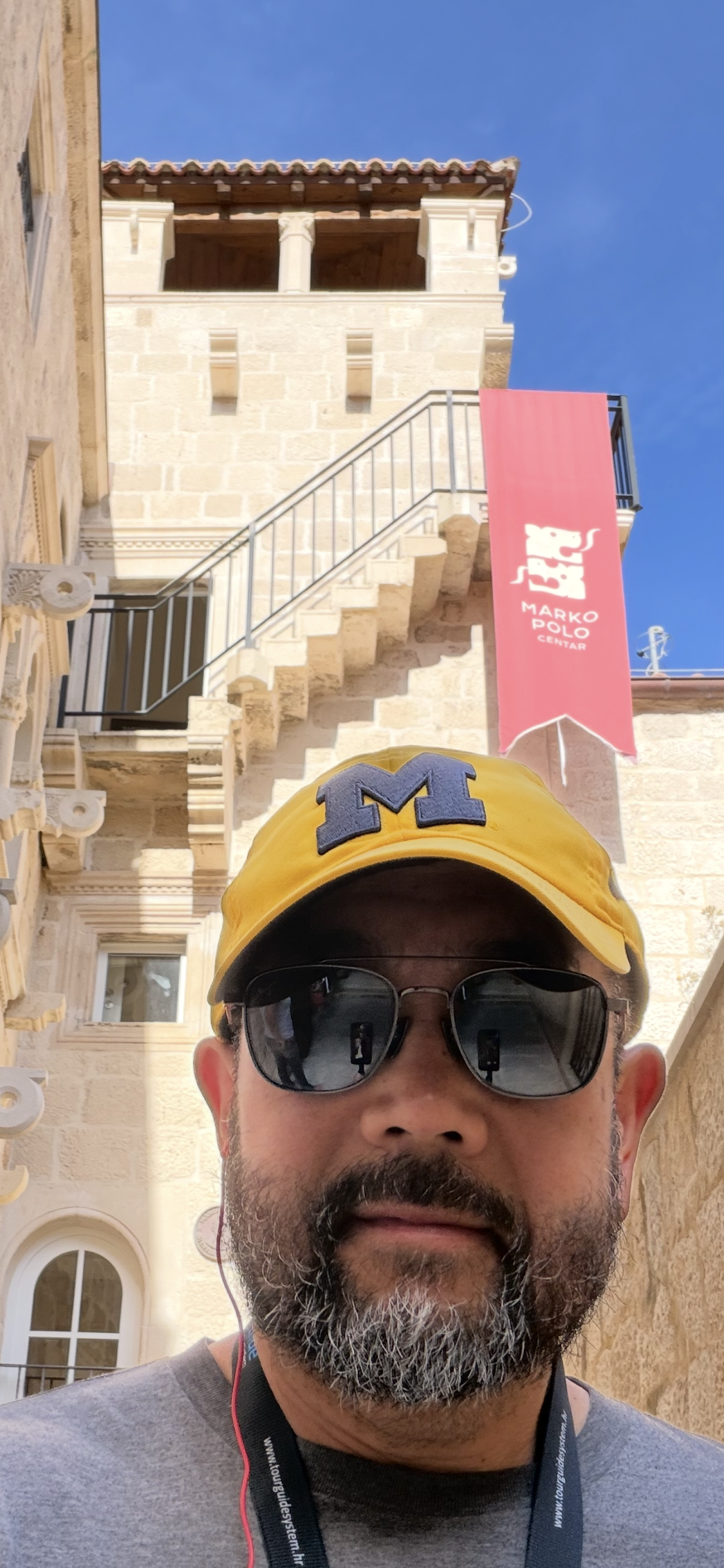Person in sunglasses and maize “M” cap stands outside a stone building with a red “Marko Polo” banner.