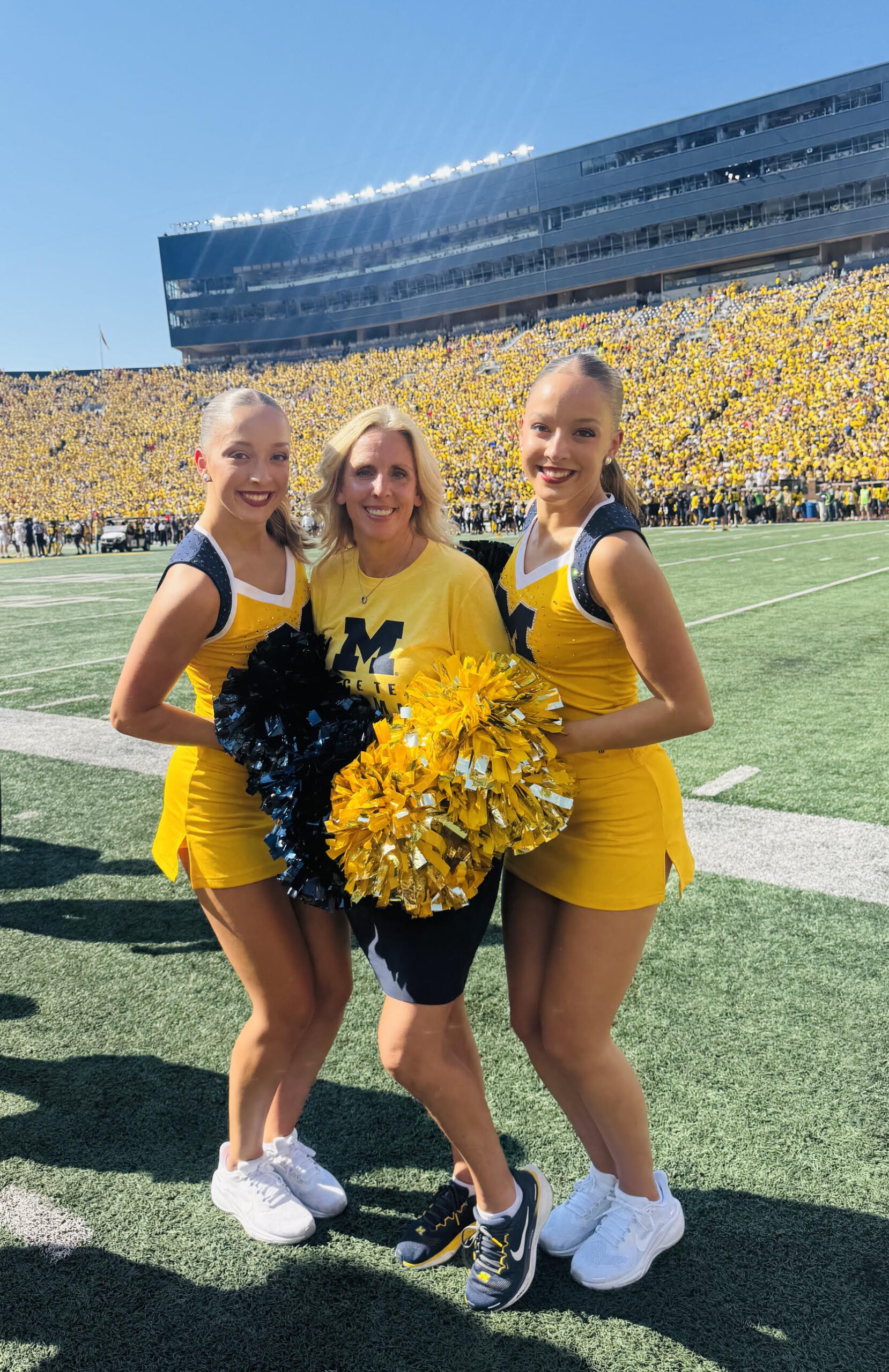 Three people in yellow cheer uniforms pose with pom-poms on a football field, with a stadium crowd in the background.