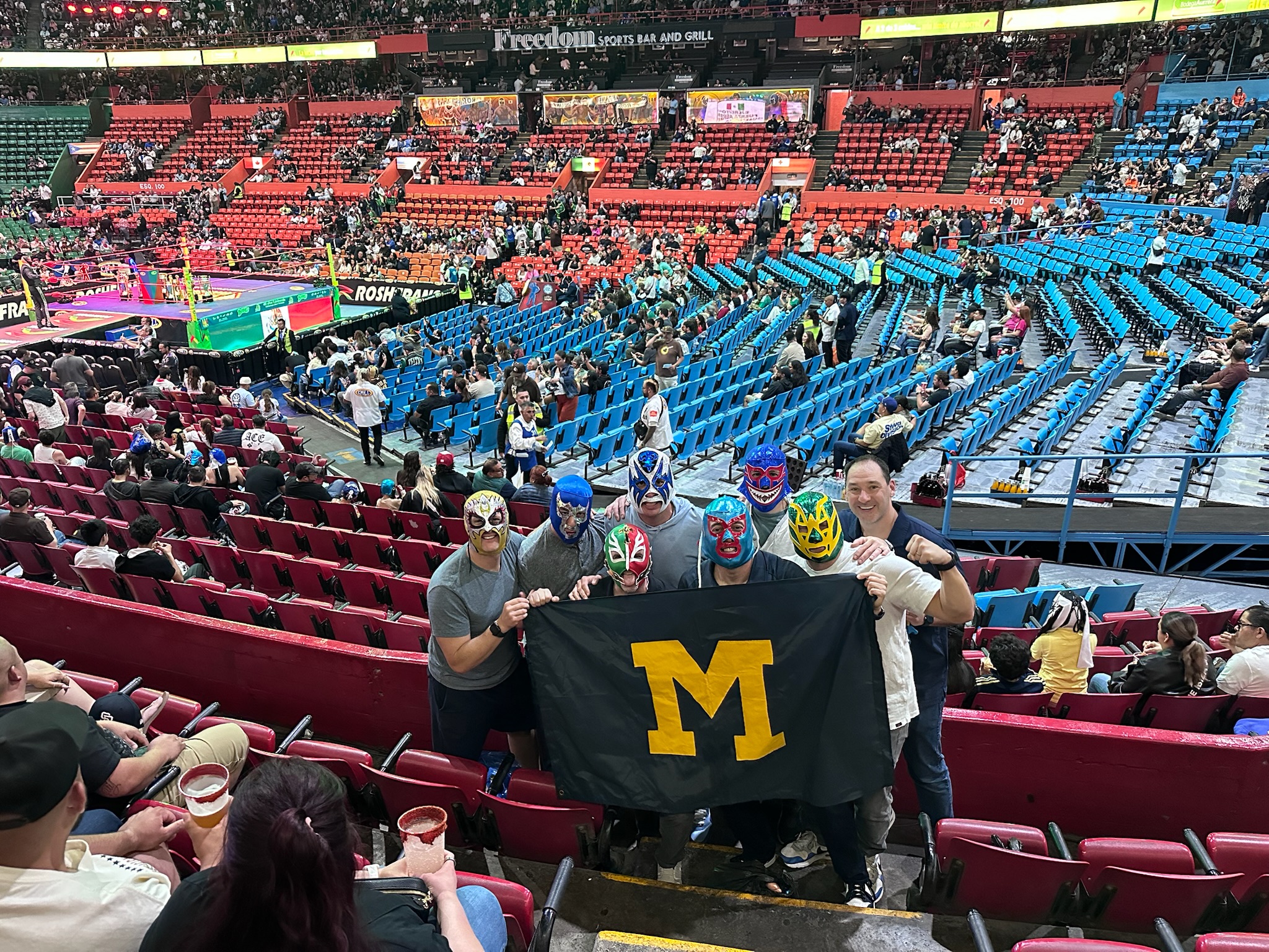 Five people in wrestling masks hold a black flag with a maize “M” at an indoor sports arena with a large crowd.
