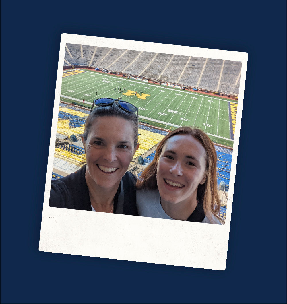 Two people smile for a selfie in front of the Michigan Stadium field, with maize and blue seating.