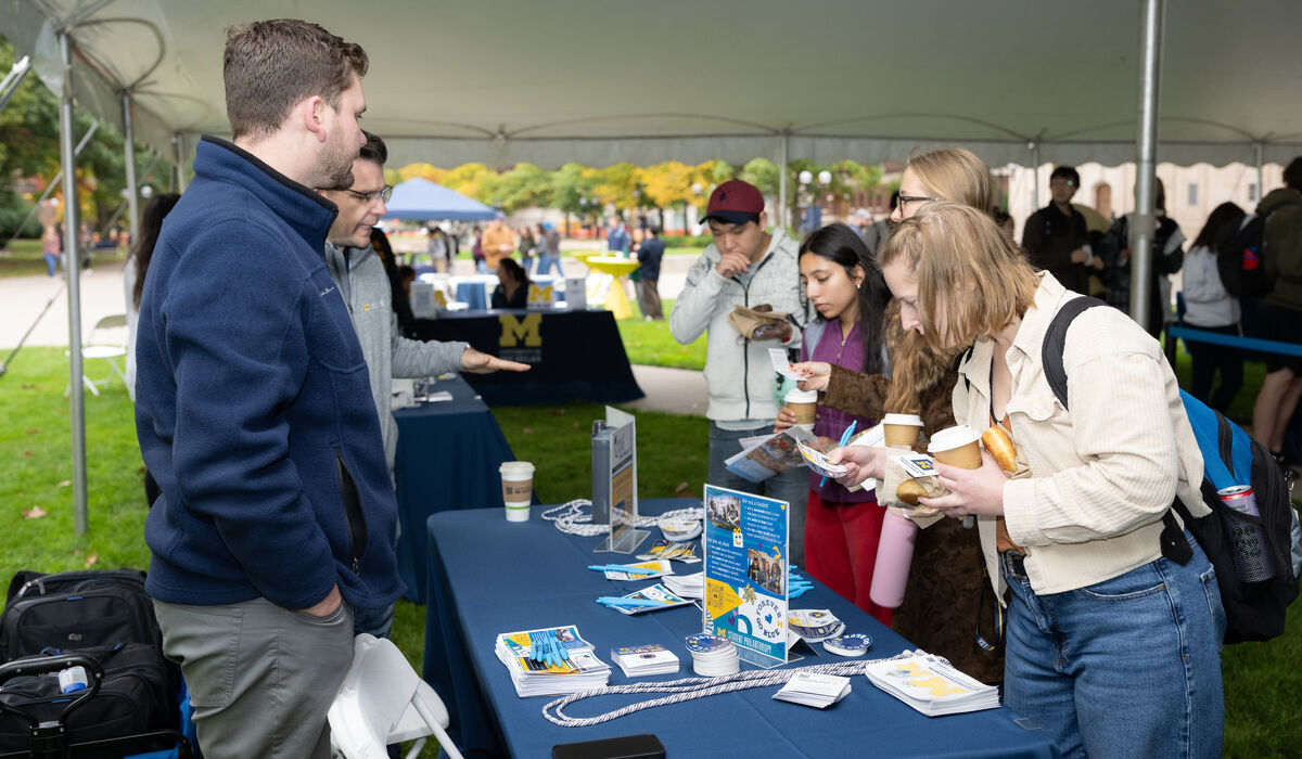 Under a white tent, individuals engage with a booth displaying brochures and coffee cups, while others browse in the background.