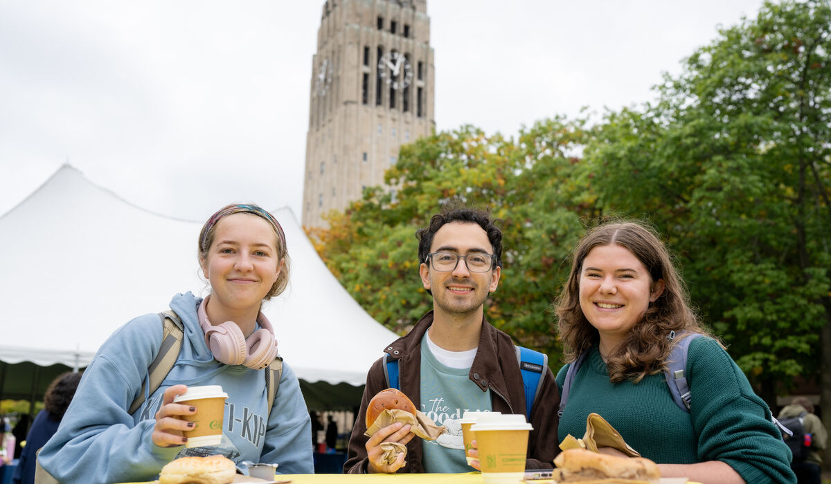 Three individuals enjoy food and drinks at a yellow table outdoors, with a clock tower and trees visible in the background.