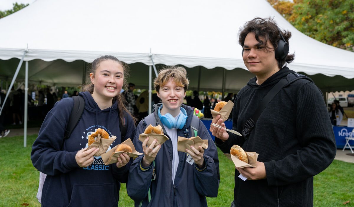 Three young people stand outdoors under a white tent, smiling and holding food items in paper wrappers.