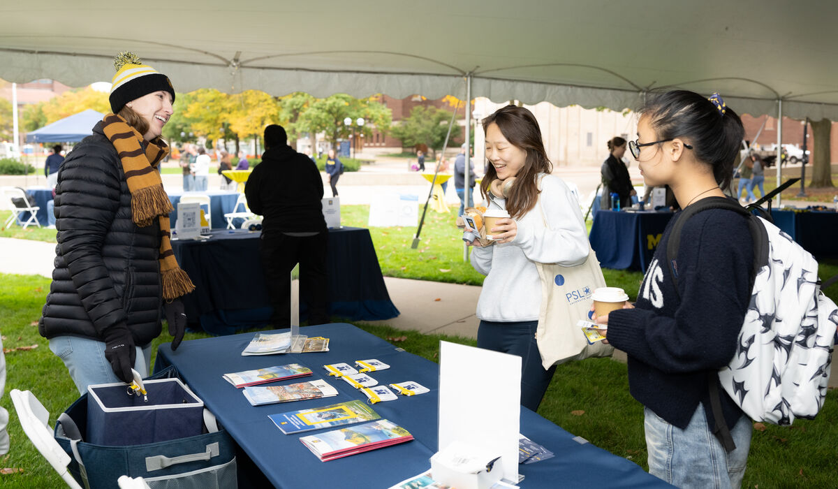 Three individuals engage in conversation under a tent at an outdoor event, with one person holding a coffee cup and another smiling while standing near a table displaying brochures and materials.