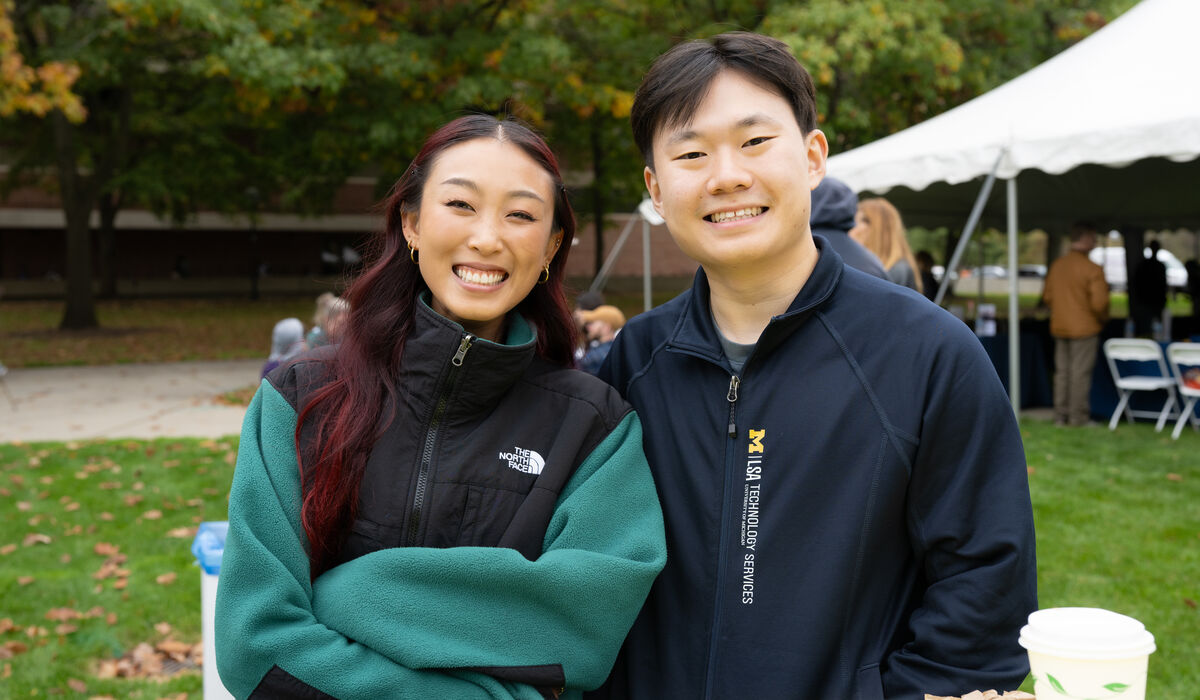 Two individuals smile for the camera while seated at an outdoor table, with autumn trees and a white tent visible in the background.