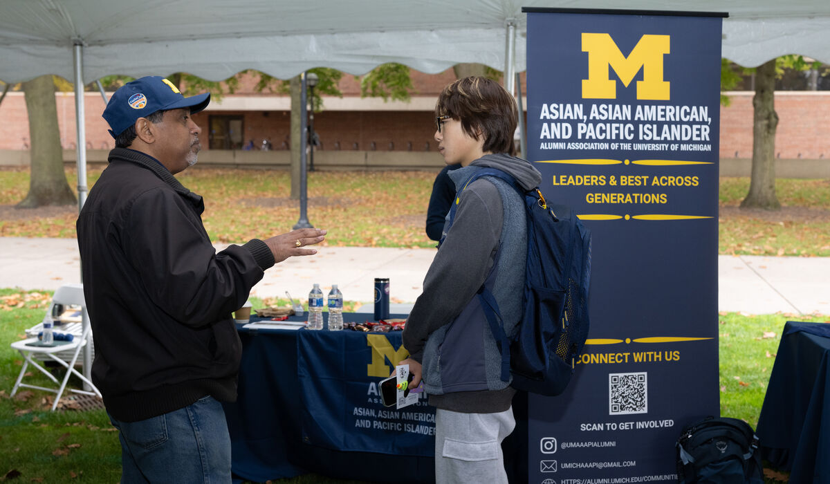 Two individuals converse under a tent at an outdoor event, with a banner for the Asian, Asian American, and Pacific Islander Alumni Association of the University of Michigan.
