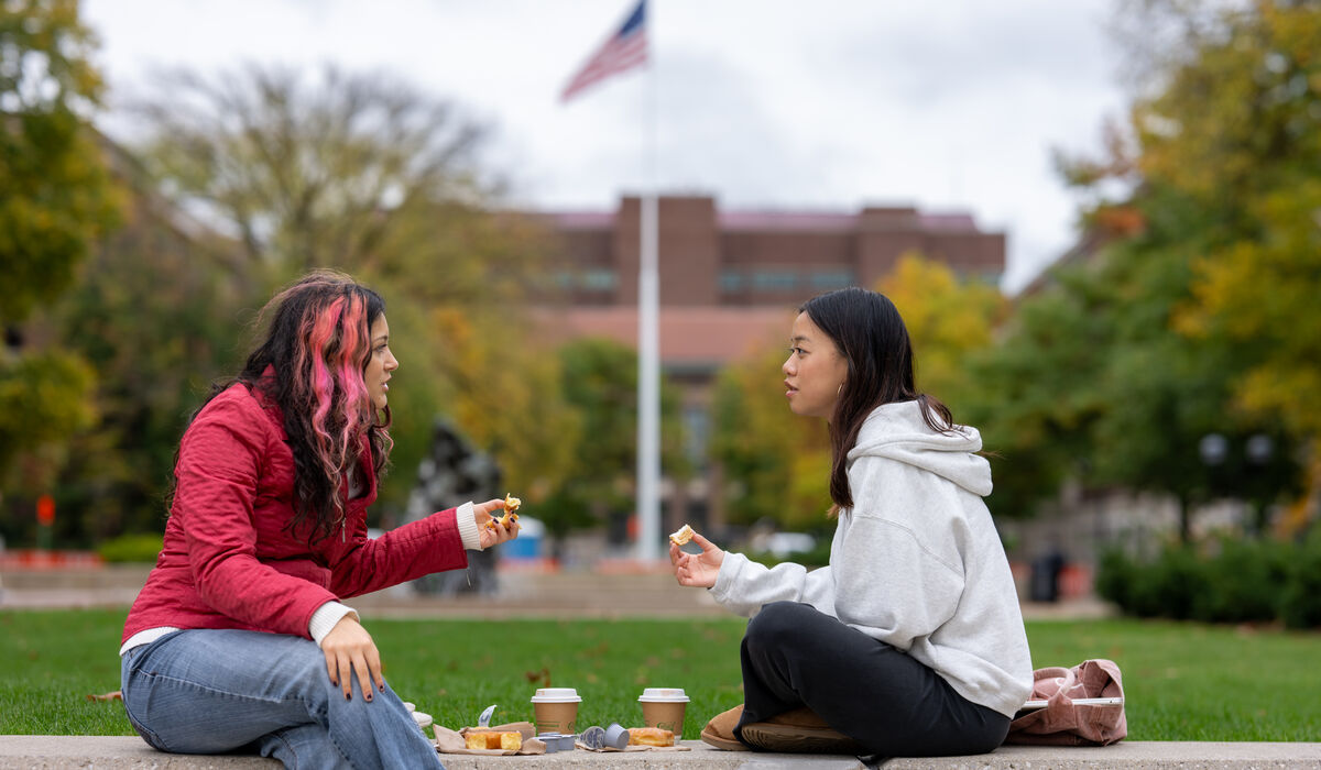 Two individuals sit on a concrete ledge outdoors, sharing snacks and drinks while conversing, with an American flag and autumn trees in the background.