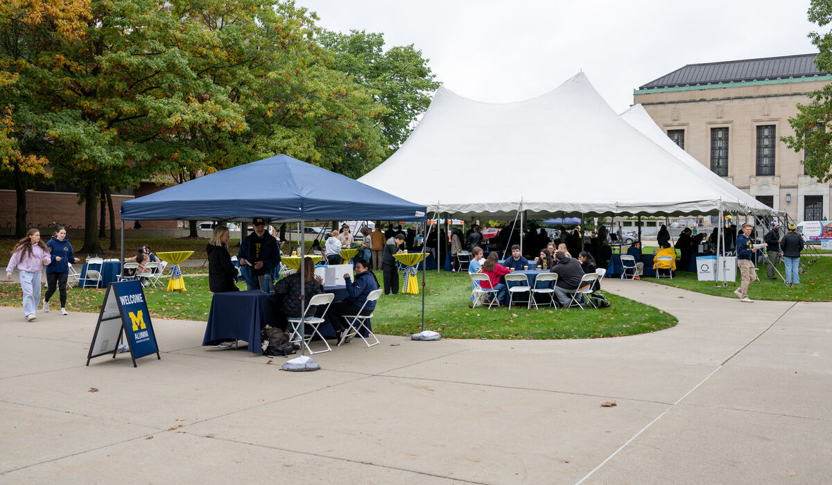 A campus event features a blue canopy with a "Welcome Michigan" sign and a large white tent where people gather on a grassy area near a building.