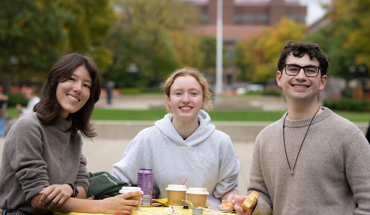 Three individuals sit at a yellow table outdoors, smiling for the camera while enjoying drinks and snacks.