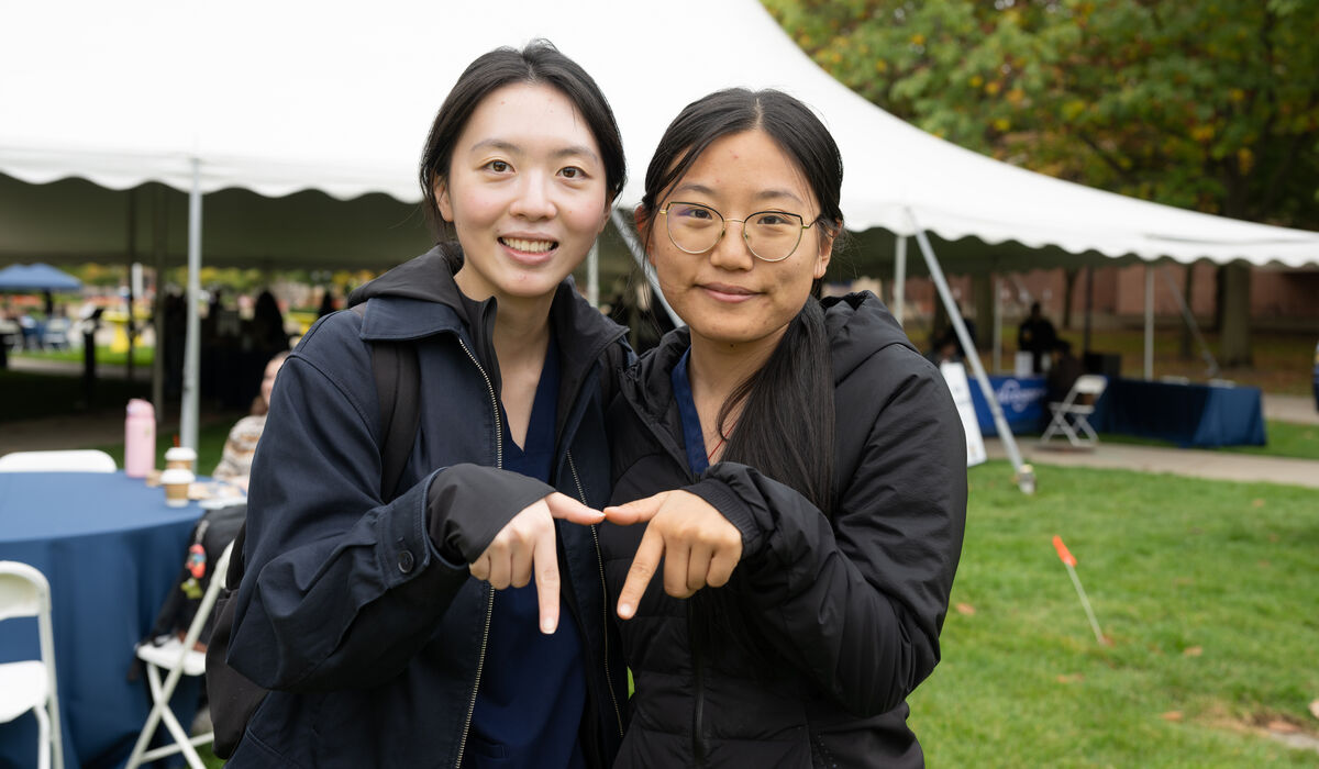 Two individuals pose together outdoors at an event, making a heart shape with their hands while smiling.