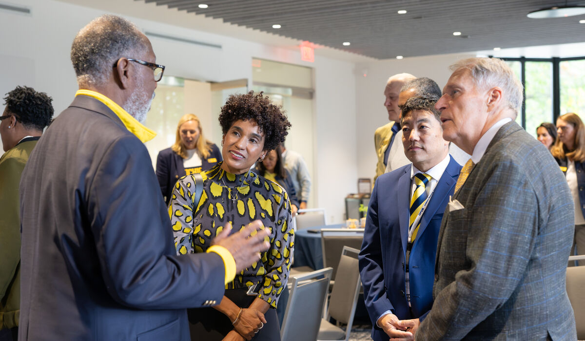 A group of formally dressed individuals engages in conversation in a modern, well-lit indoor space with rows of chairs.