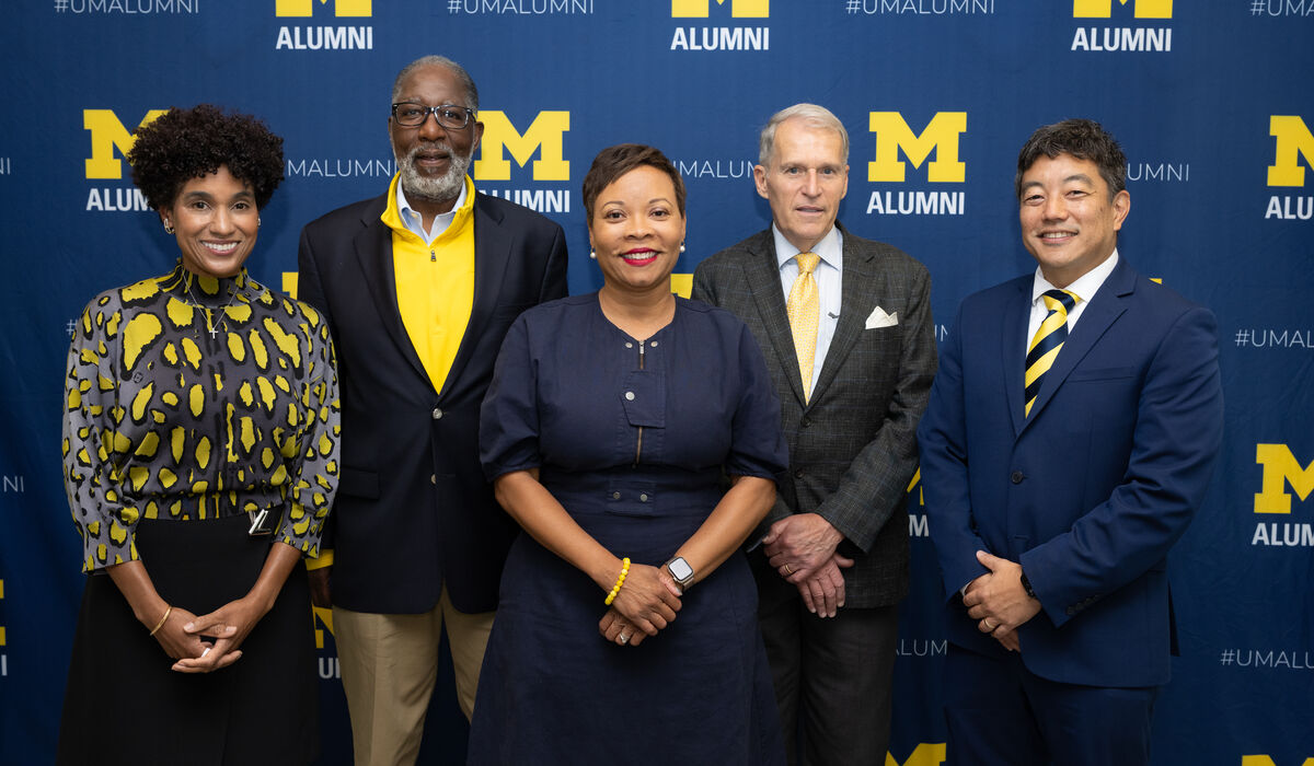 Five individuals pose together in front of a blue backdrop featuring the University of Michigan Alumni logo and hashtag.