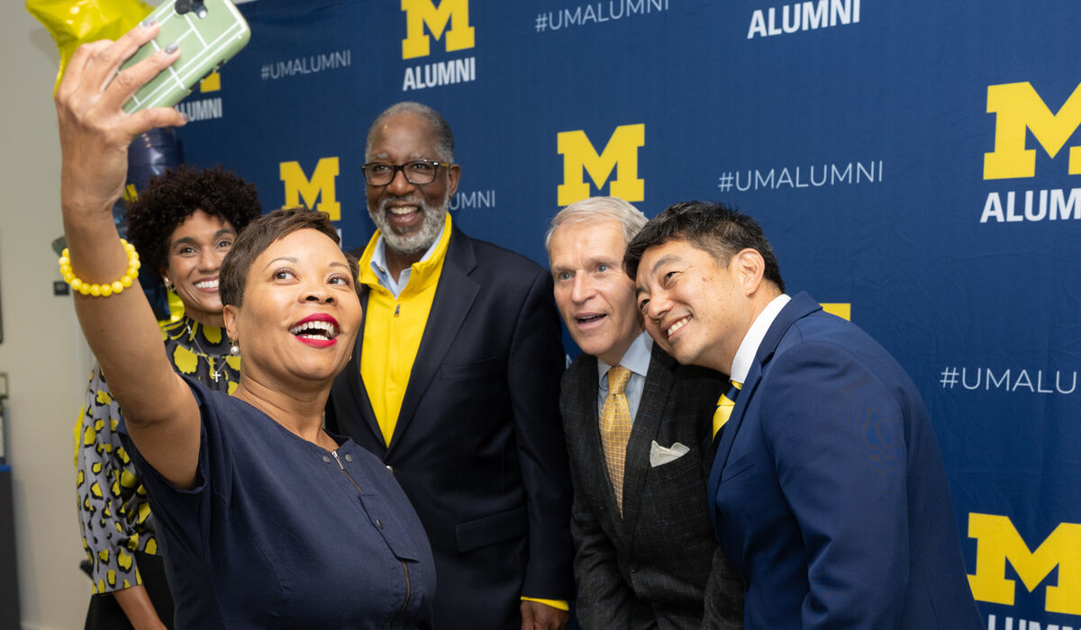 A group of five individuals poses for a selfie in front of a blue backdrop featuring the University of Michigan alumni logo and hashtag.