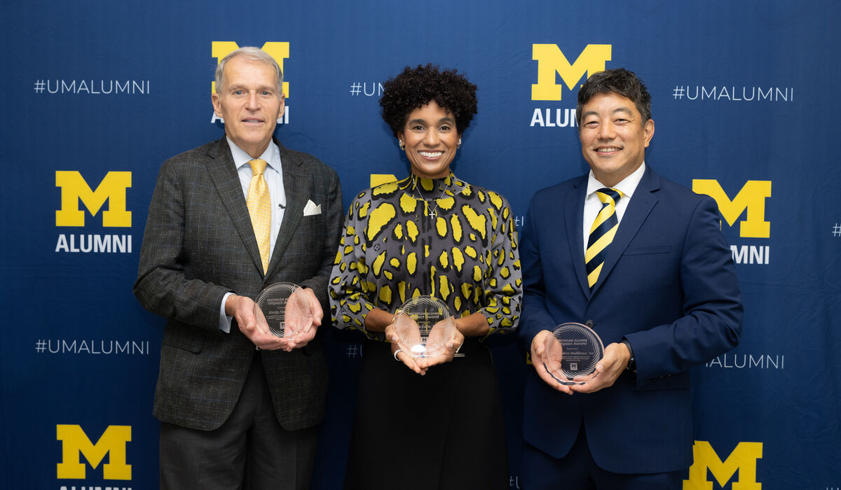 Three individuals stand together in front of a blue backdrop featuring the University of Michigan alumni logo, each holding a clear award.