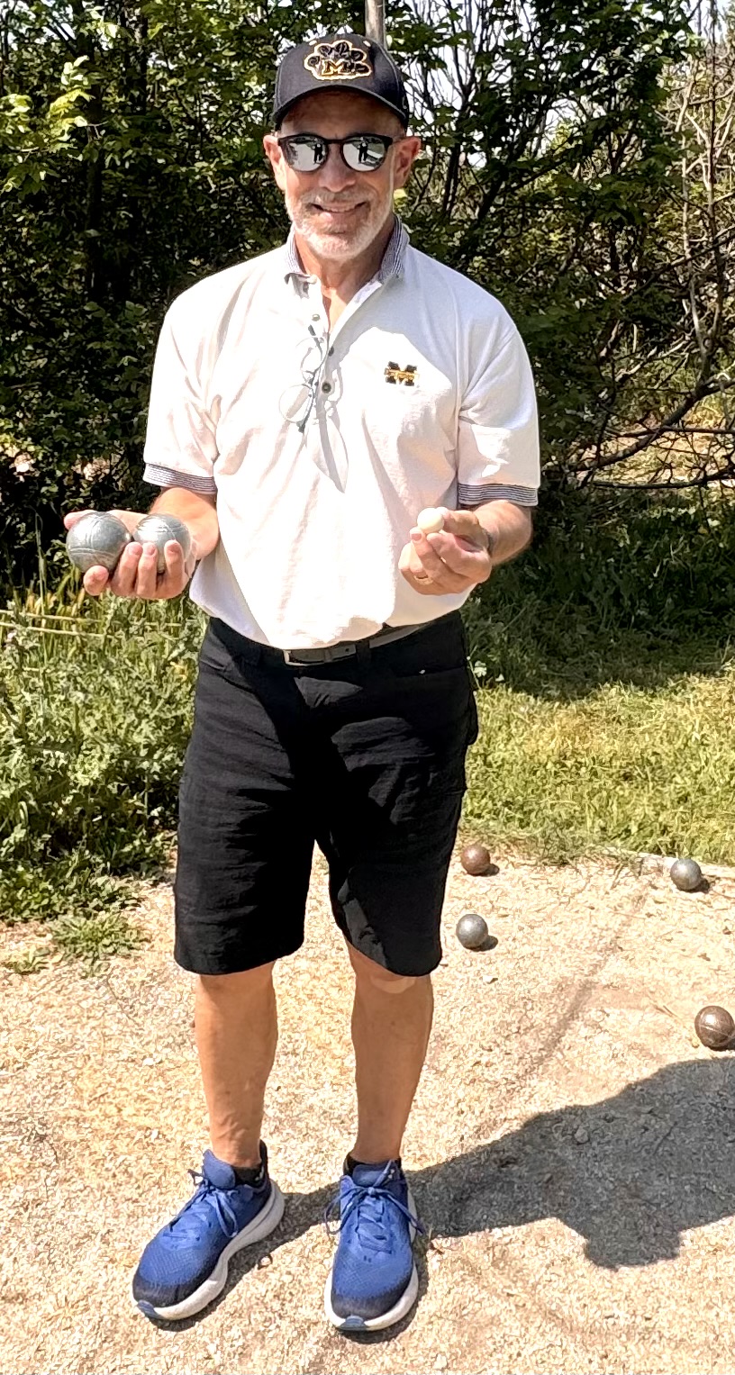 Person in sunglasses and a cap holds bocce balls, standing outdoors on a gravel court with greenery behind.