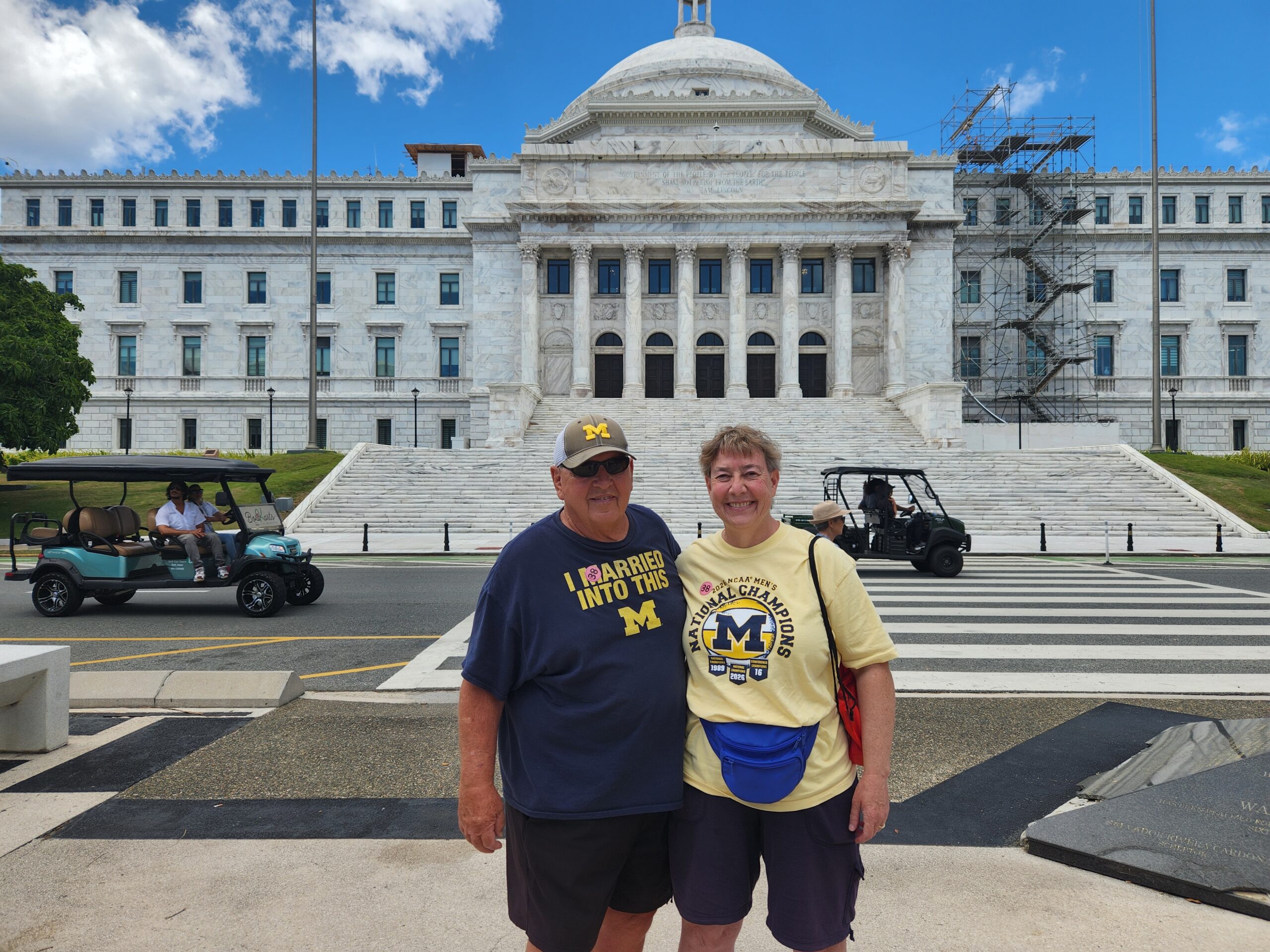 Two people in Michigan shirts pose together in front of a large white government building with columns and steps.