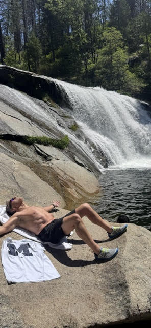 A person in shorts and sneakers lies on a towel on rocks by a waterfall, arms outstretched, in the sun.