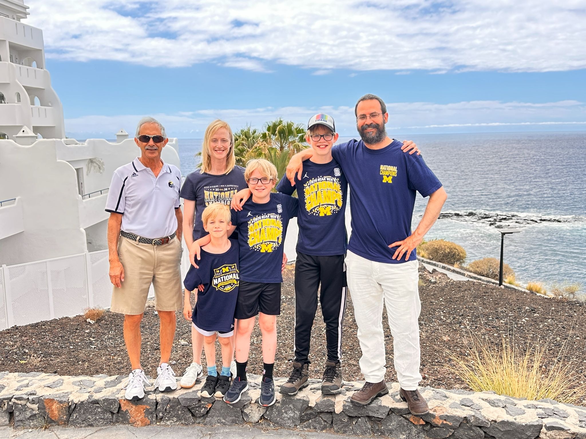 Six people pose on a stone path near the ocean, with a white building, plants, and a partly cloudy sky in the background.
