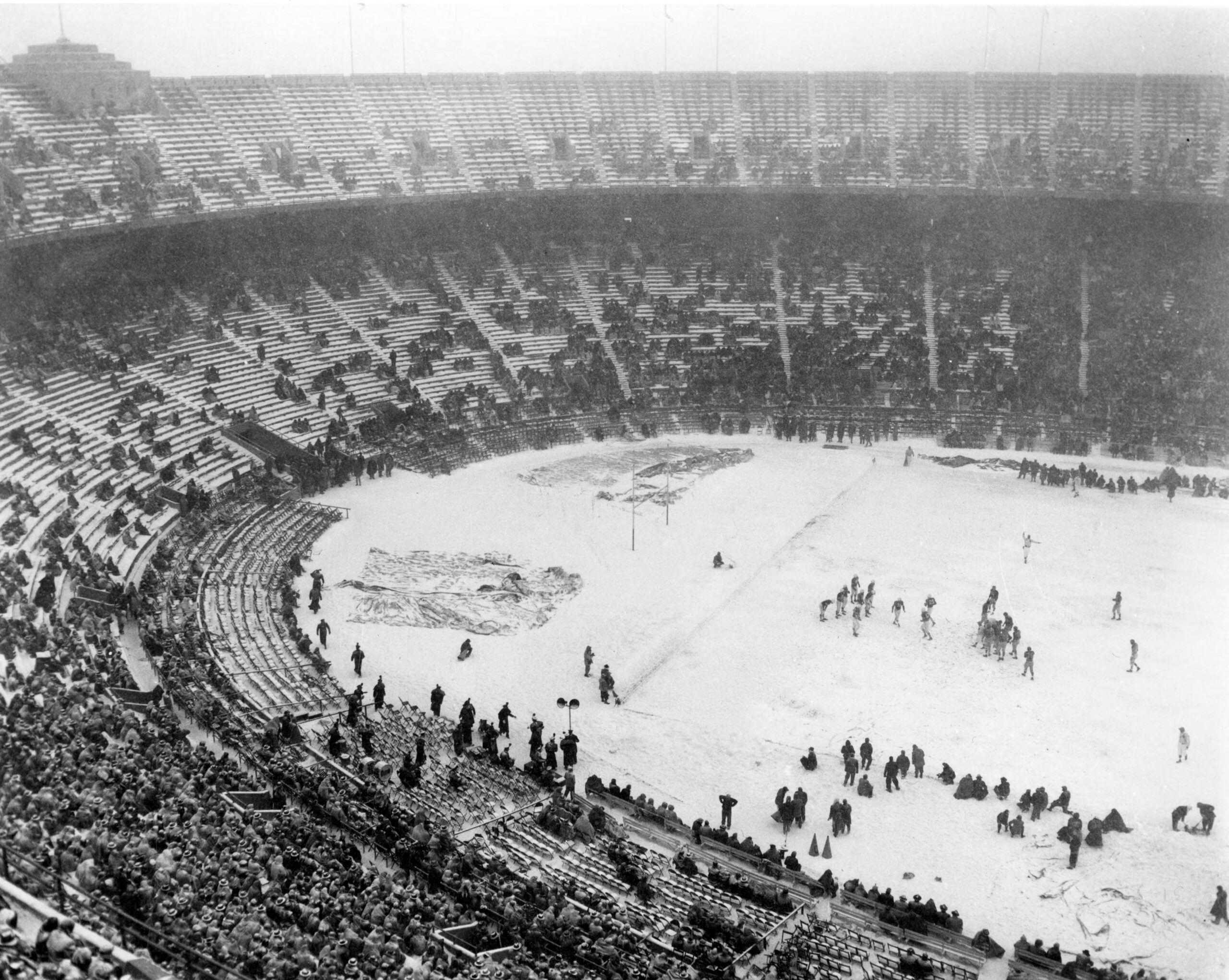 A football game takes place in a snow-covered stadium with mostly empty seats and people bundled in winter clothing.