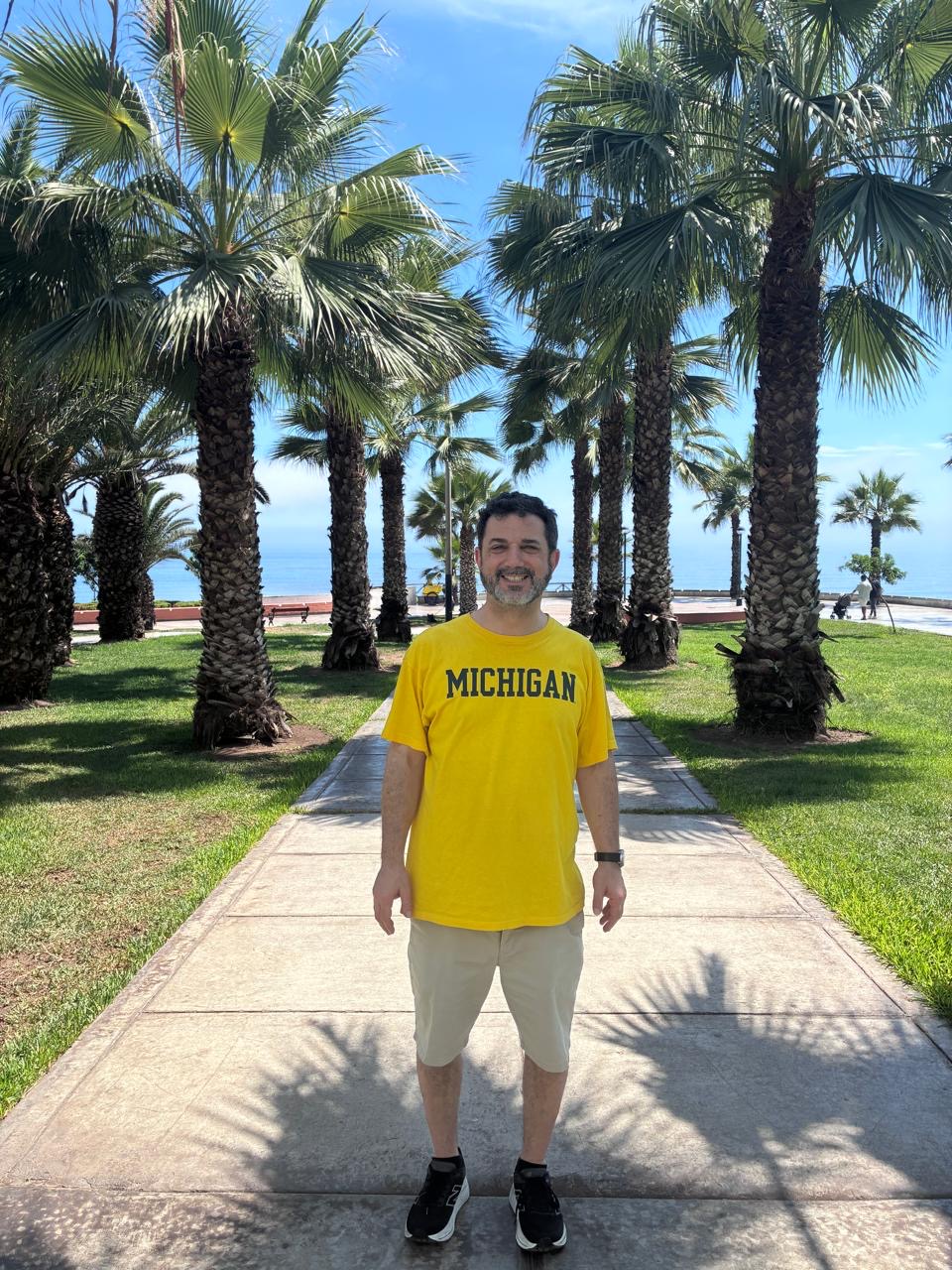 Person in a maize Michigan shirt stands on a sunny, palm-lined path, smiling at the camera.