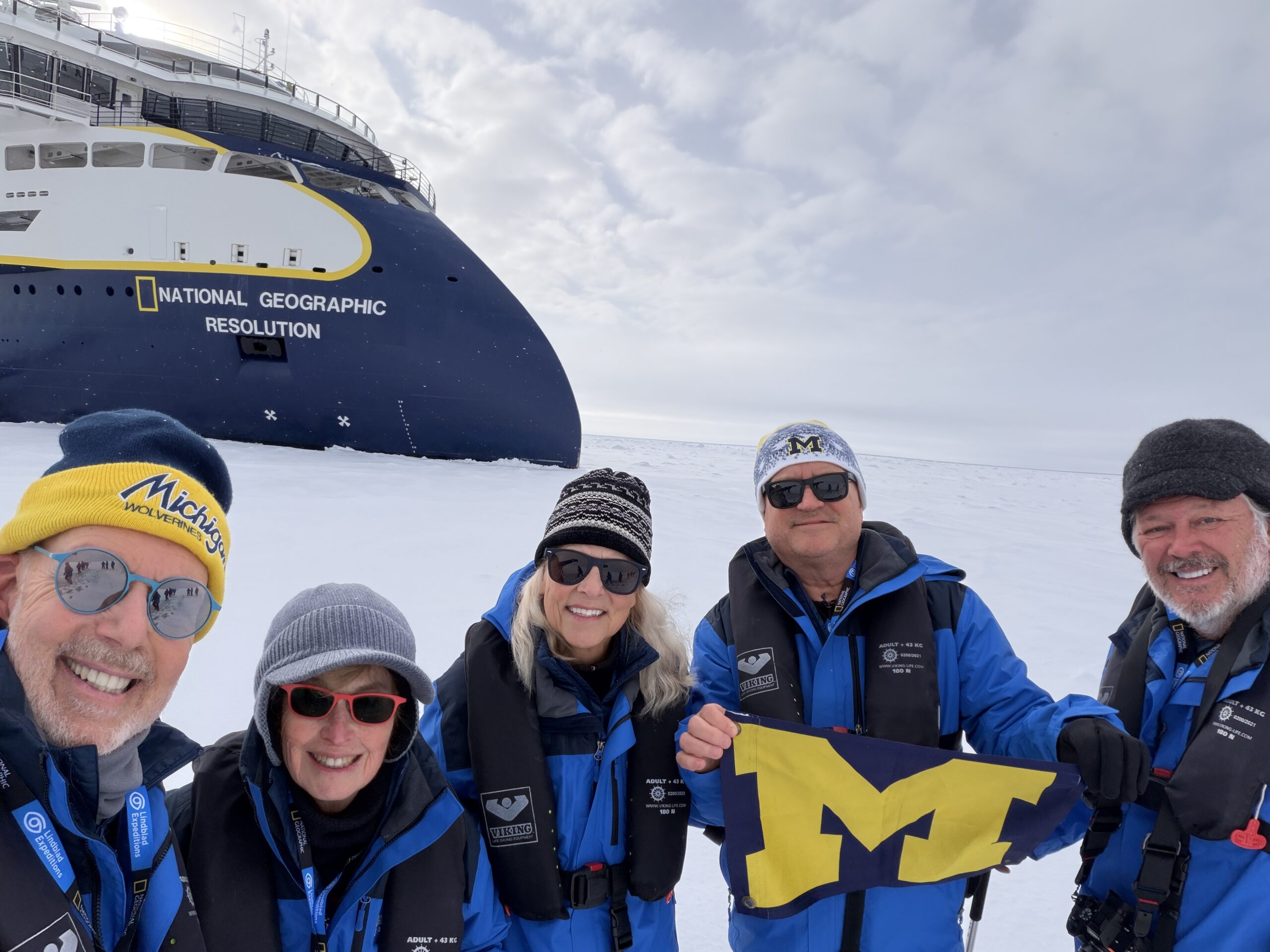 Five people in winter gear pose on snow by a National Geographic ship, holding a maize and blue Michigan flag.