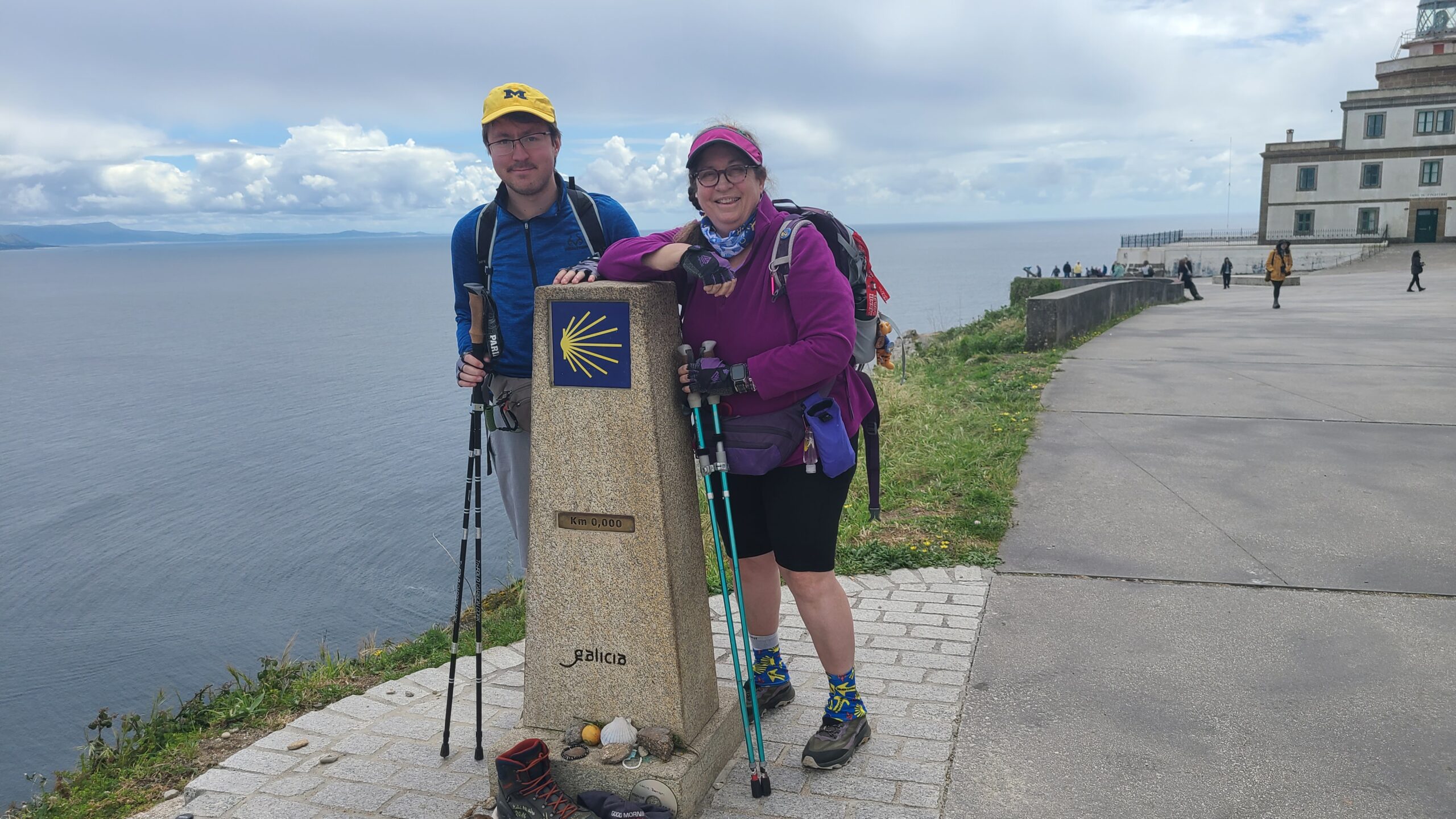 Two people with backpacks and trekking poles stand next to a Camino de Santiago marker by a coastline and a building.