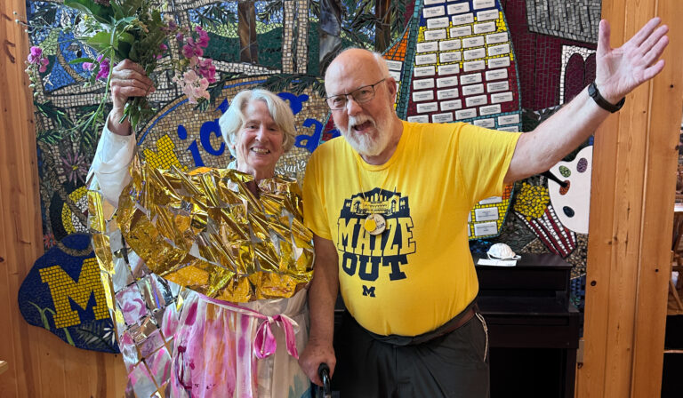 Two people smile indoors; one holds flowers and wears shiny gold material, the other wears a maize “Maize Out” shirt.