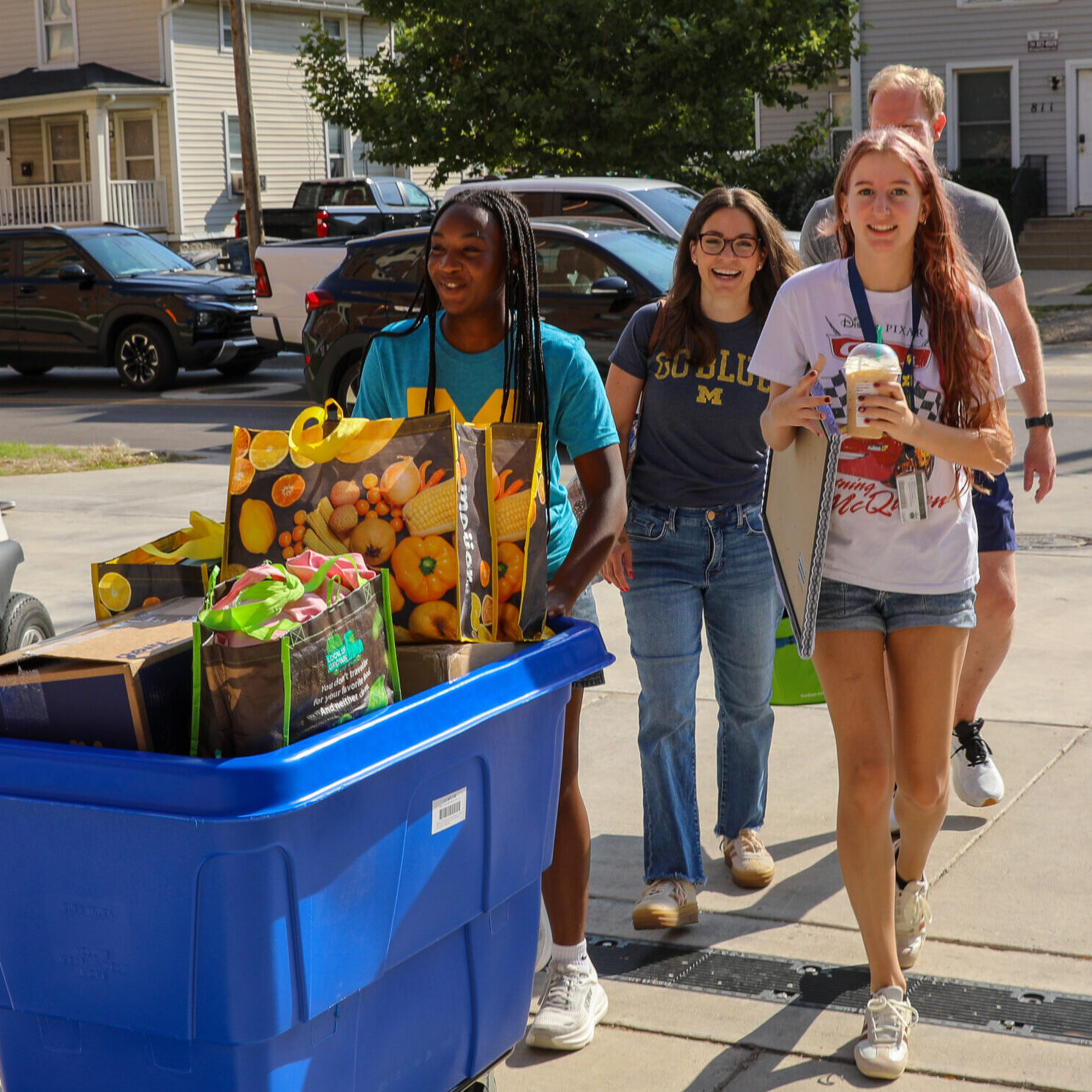 Three people push a blue bin filled with boxes and bags down a sidewalk on a sunny day.