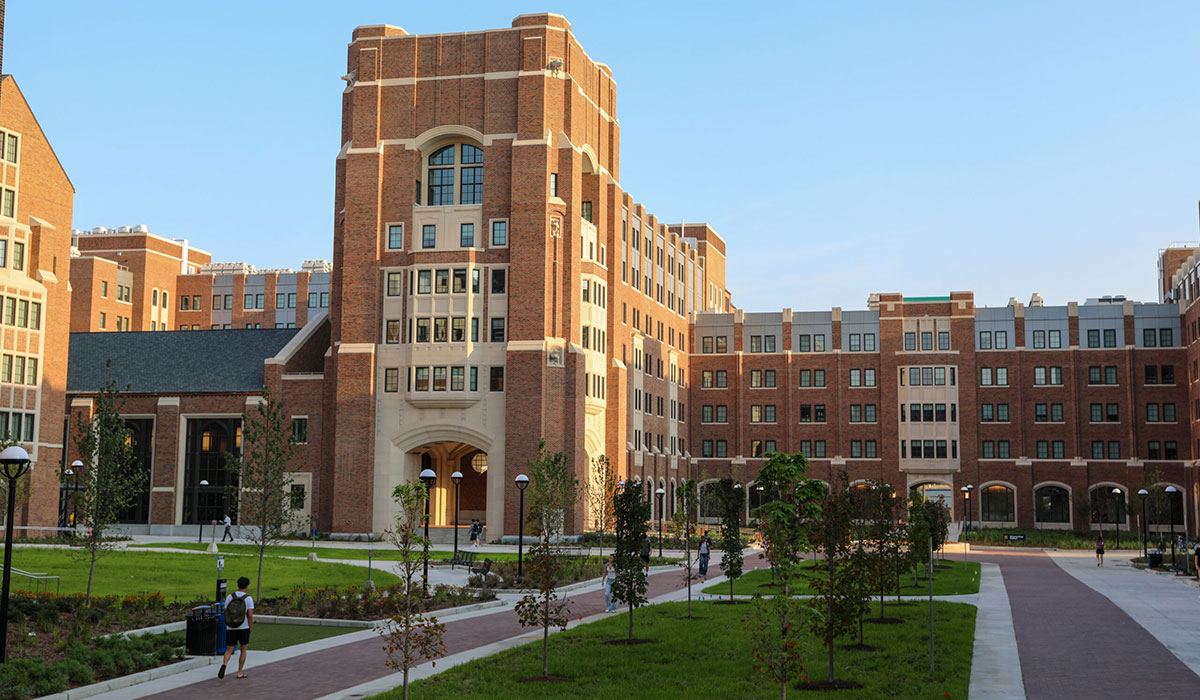 A large brick university building with many windows, an archway entrance, and walkways bordered by green lawns.