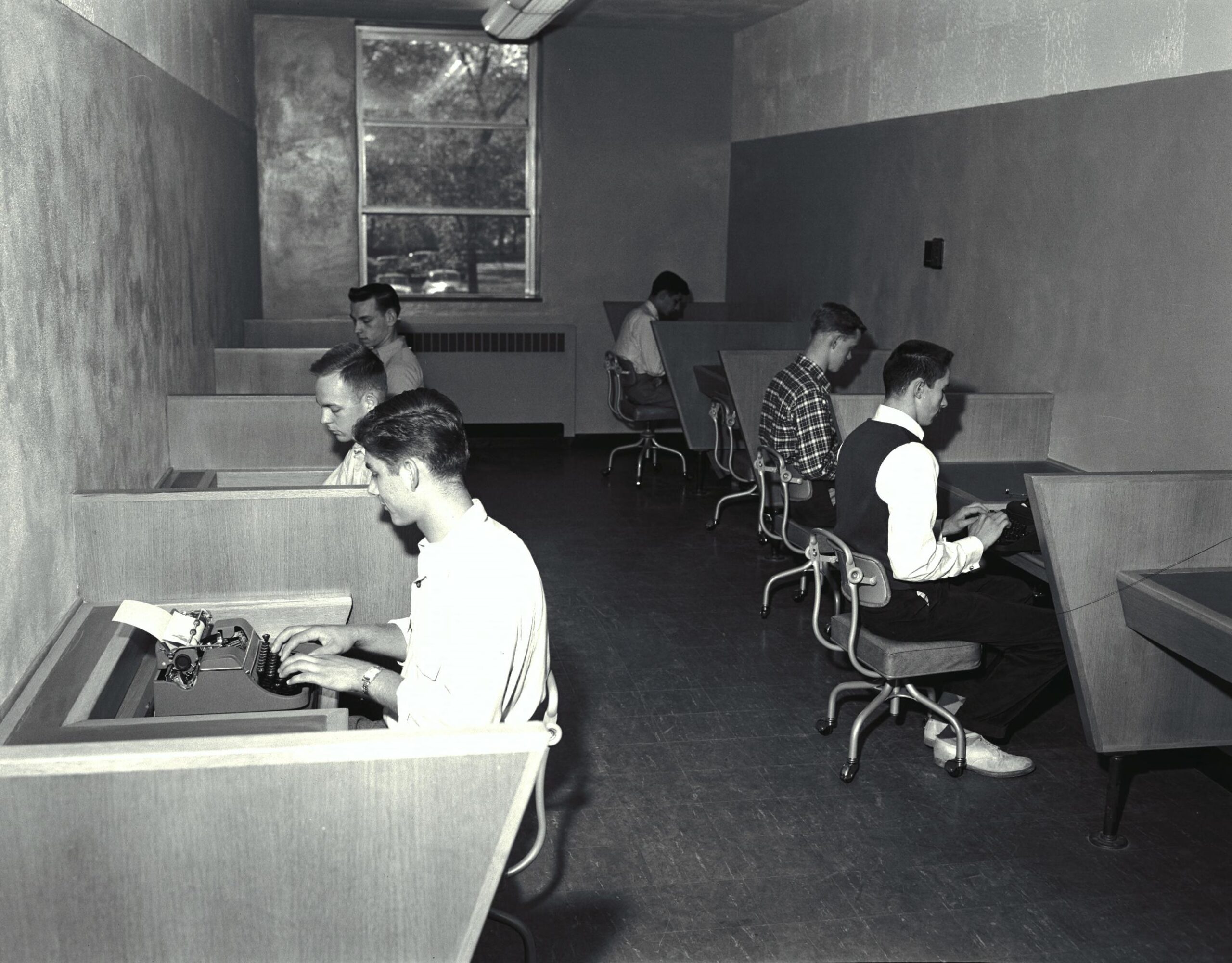 Six people sit at desks in a small room, each typing at a typewriter, with a window providing natural light at the back.
