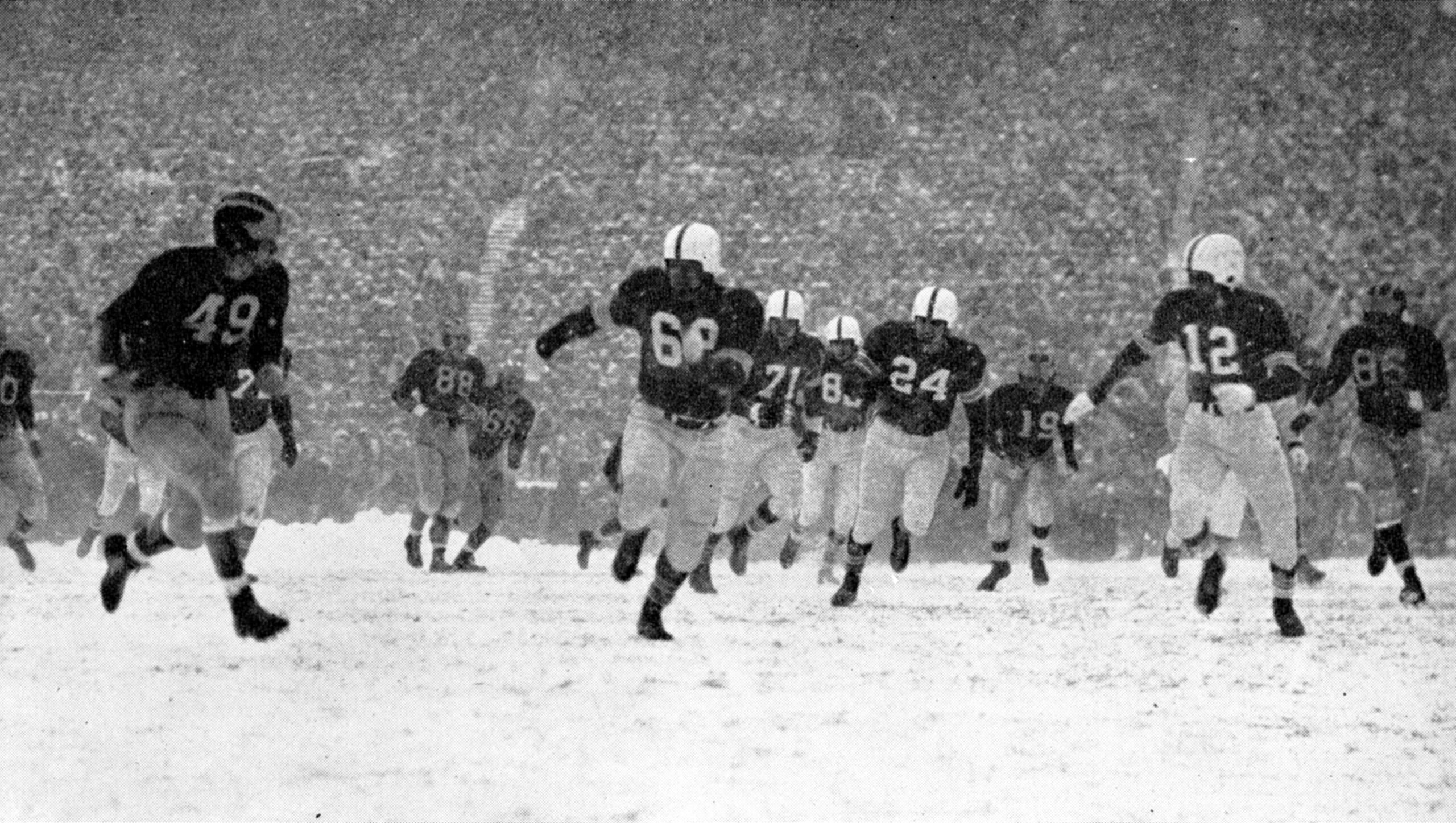 Football players run on a snow-covered field during a game, with fans visible in the background.