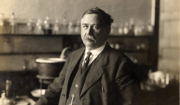 A person in a suit stands in a laboratory with glassware on shelves and a table behind them.
