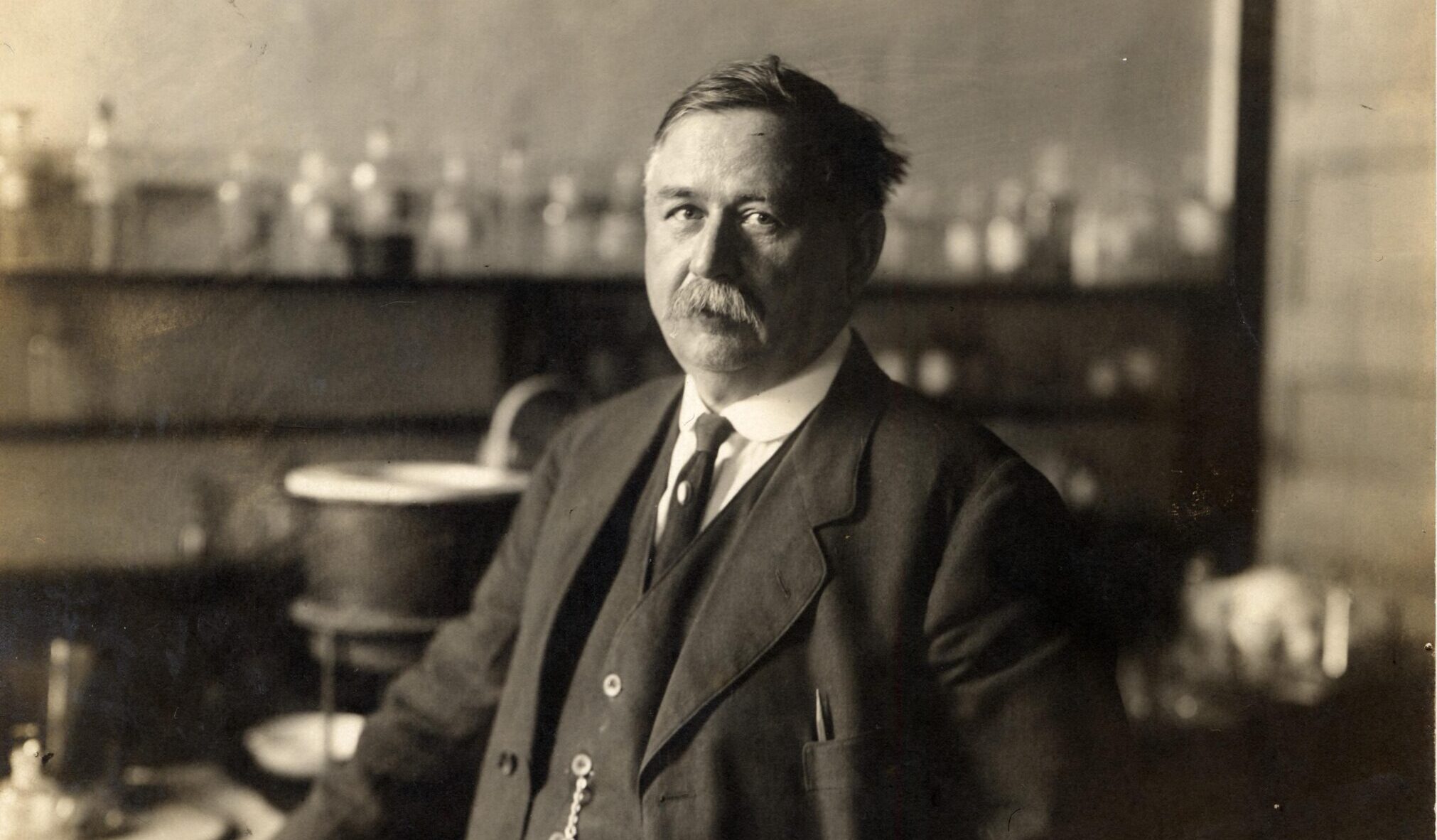 A person in a suit stands in a laboratory with glassware on shelves and a table behind them.