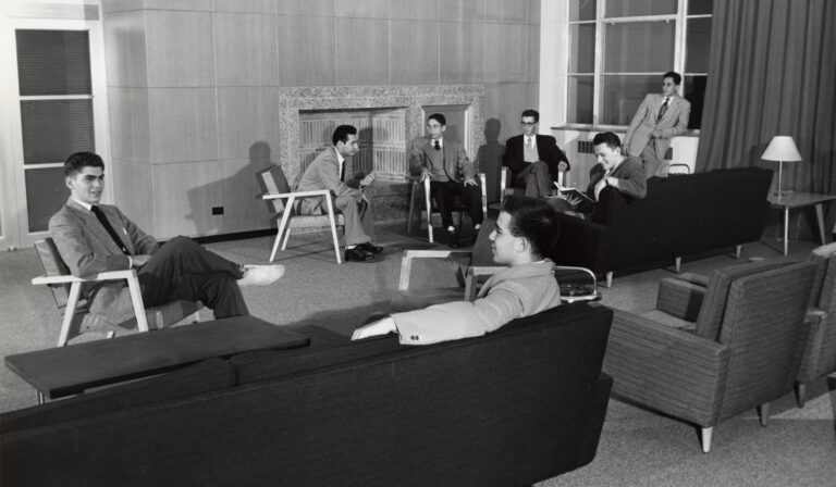 Six people in suits sit in a mid-century modern lounge, conversing near a fireplace, and large windows.