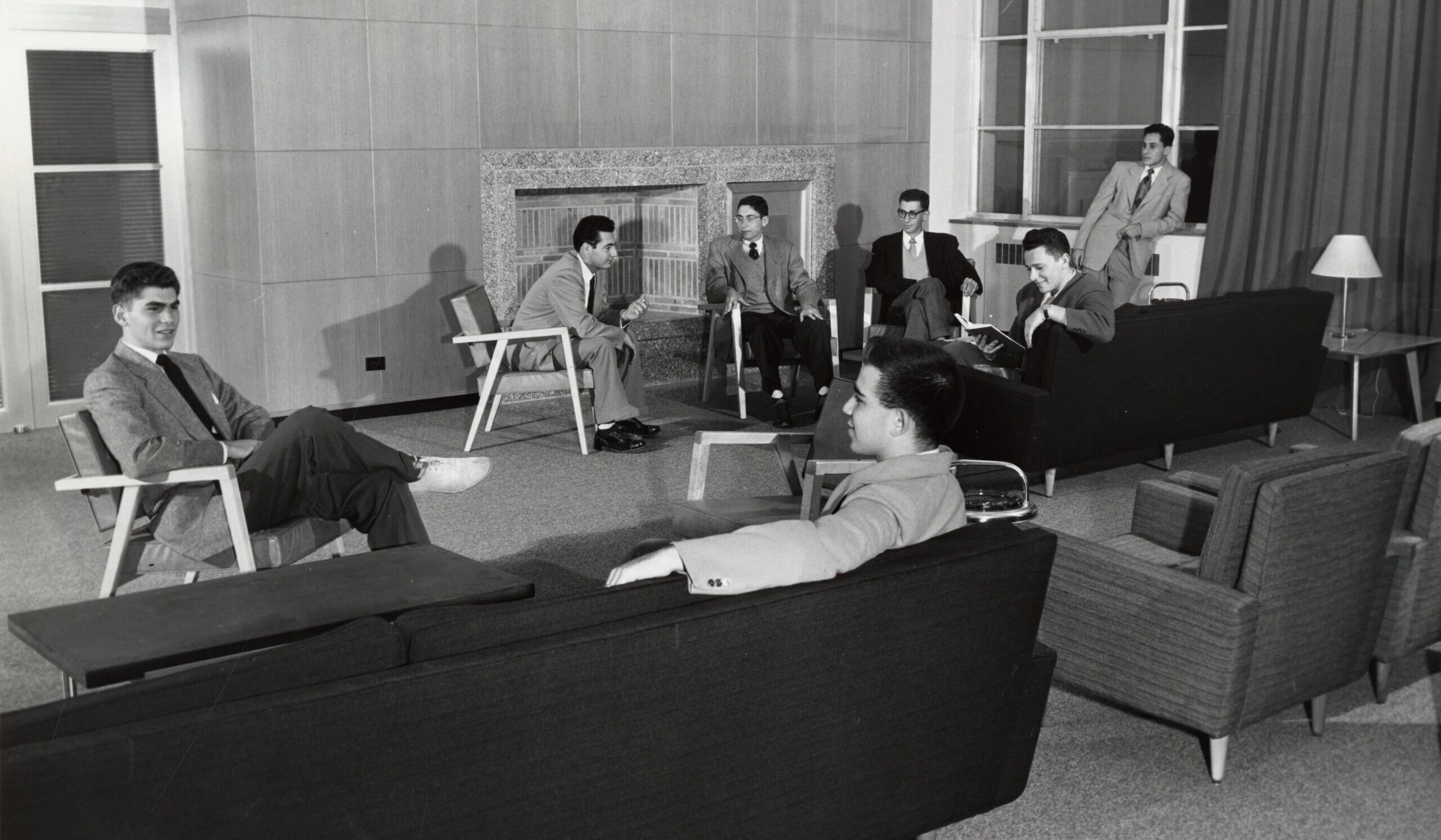 Six people in suits sit in a mid-century modern lounge, conversing near a fireplace, and large windows.