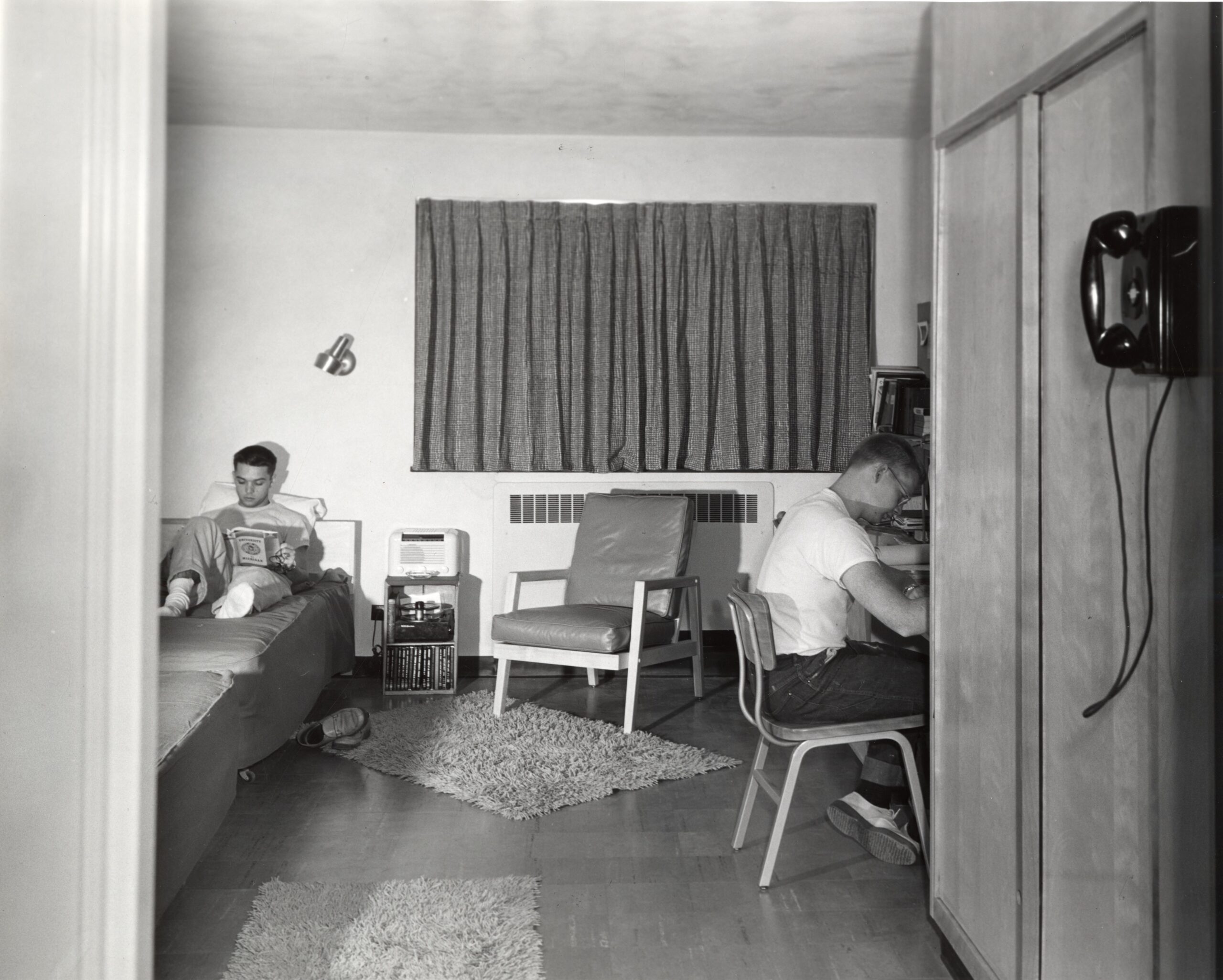 Two people in a dorm room; one reads on a bed, the other writes at a desk. Chair, phone, and rug are visible.