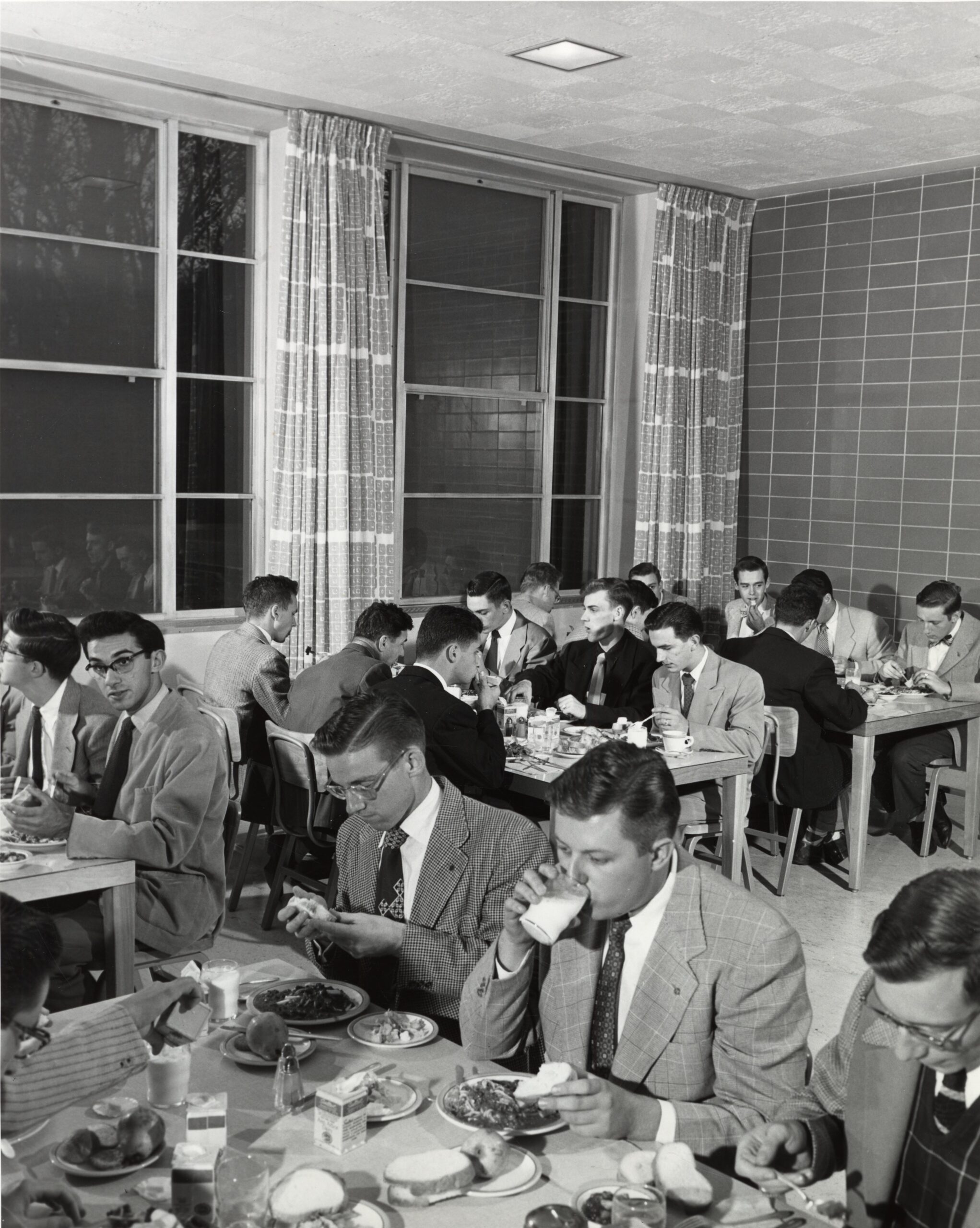 People in suits sit at tables eating lunch in a cafeteria with large windows, tiled walls, and bright lighting.